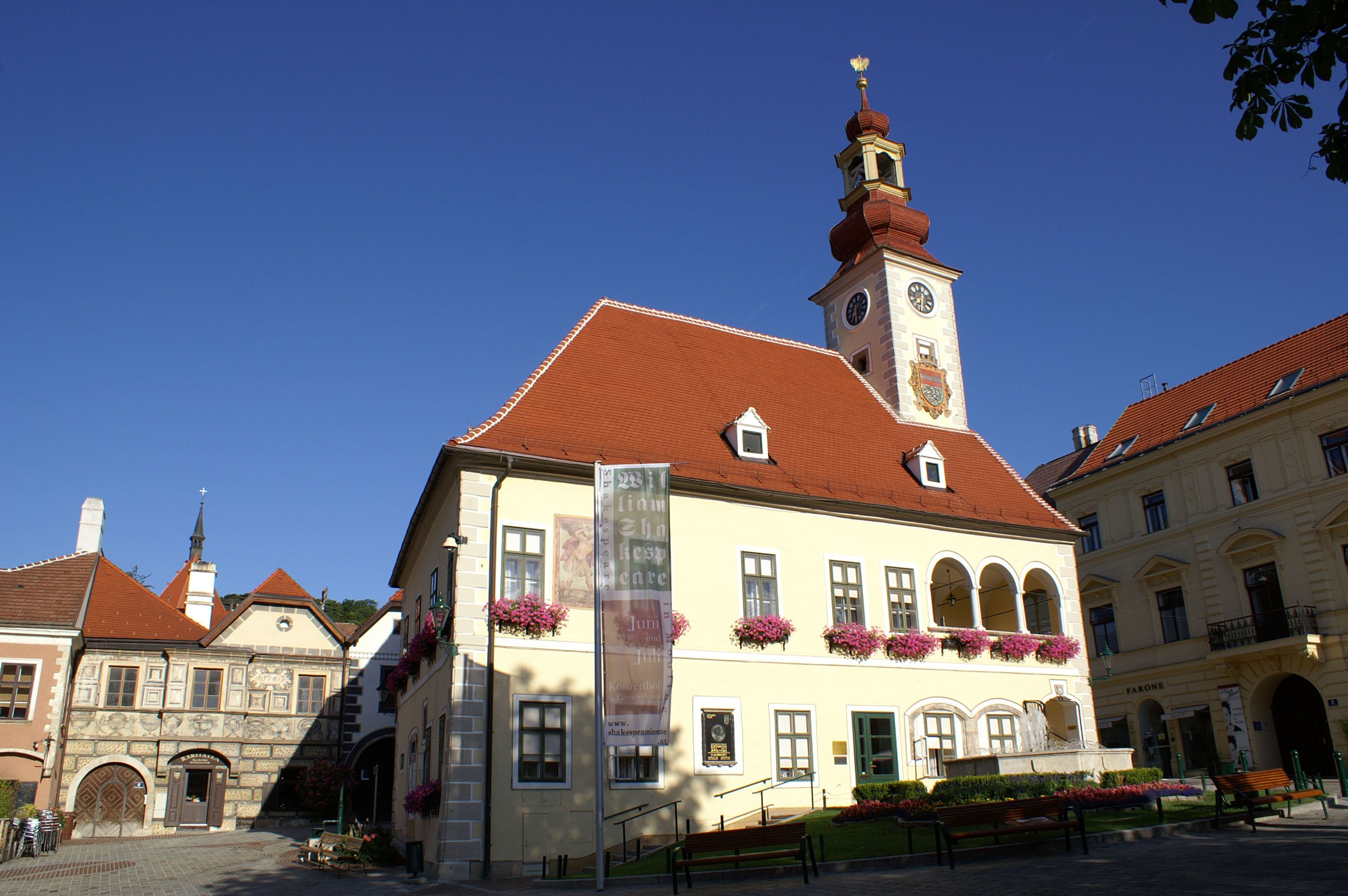 Historická budova s věží a hodinami v Mödlingu, obklopená modrou oblohou.