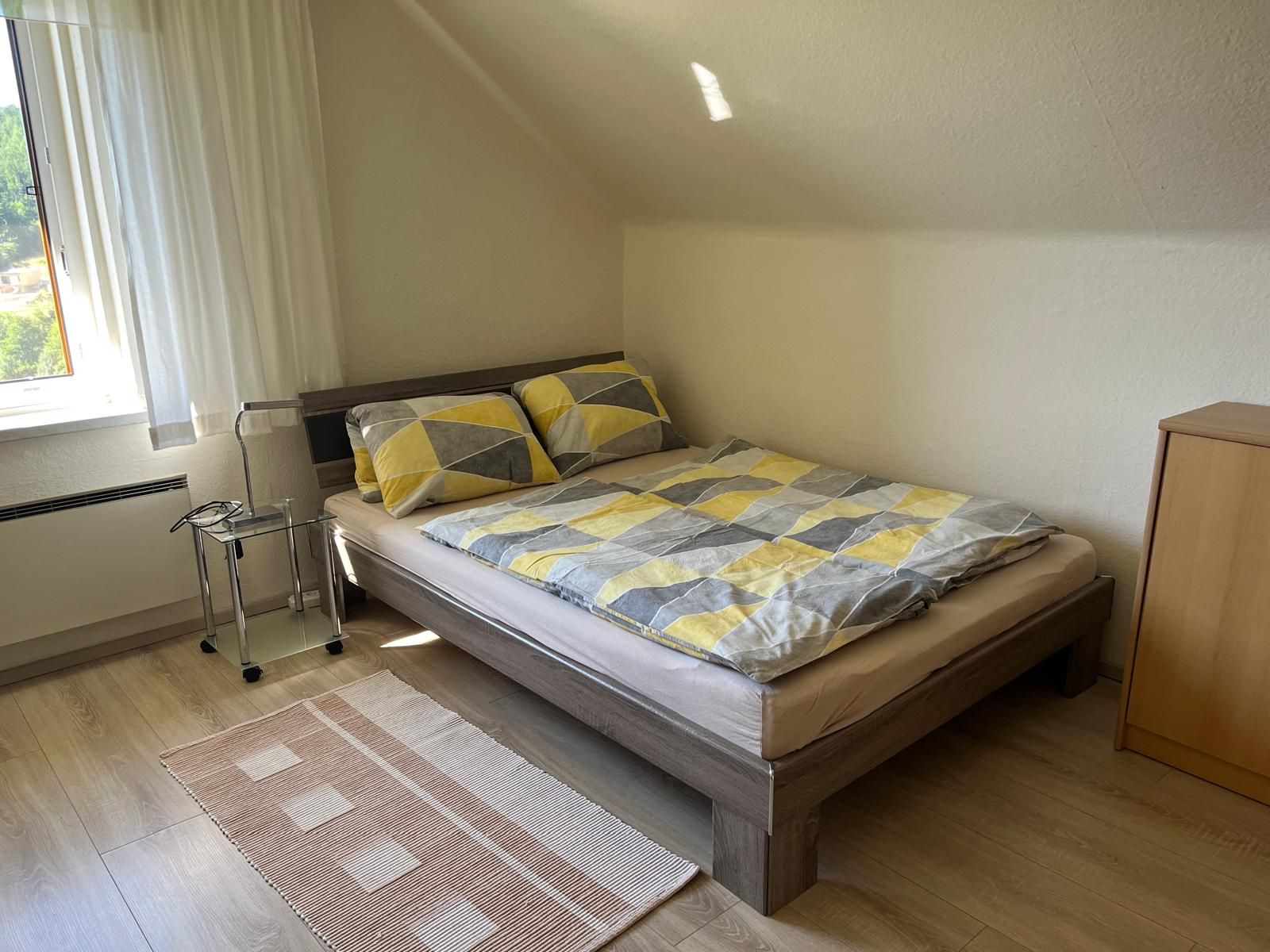 Útulná ložnice s manželskou postelí, vzorovanými peřinami, malým nočním stolkem a oknem se závěsy.