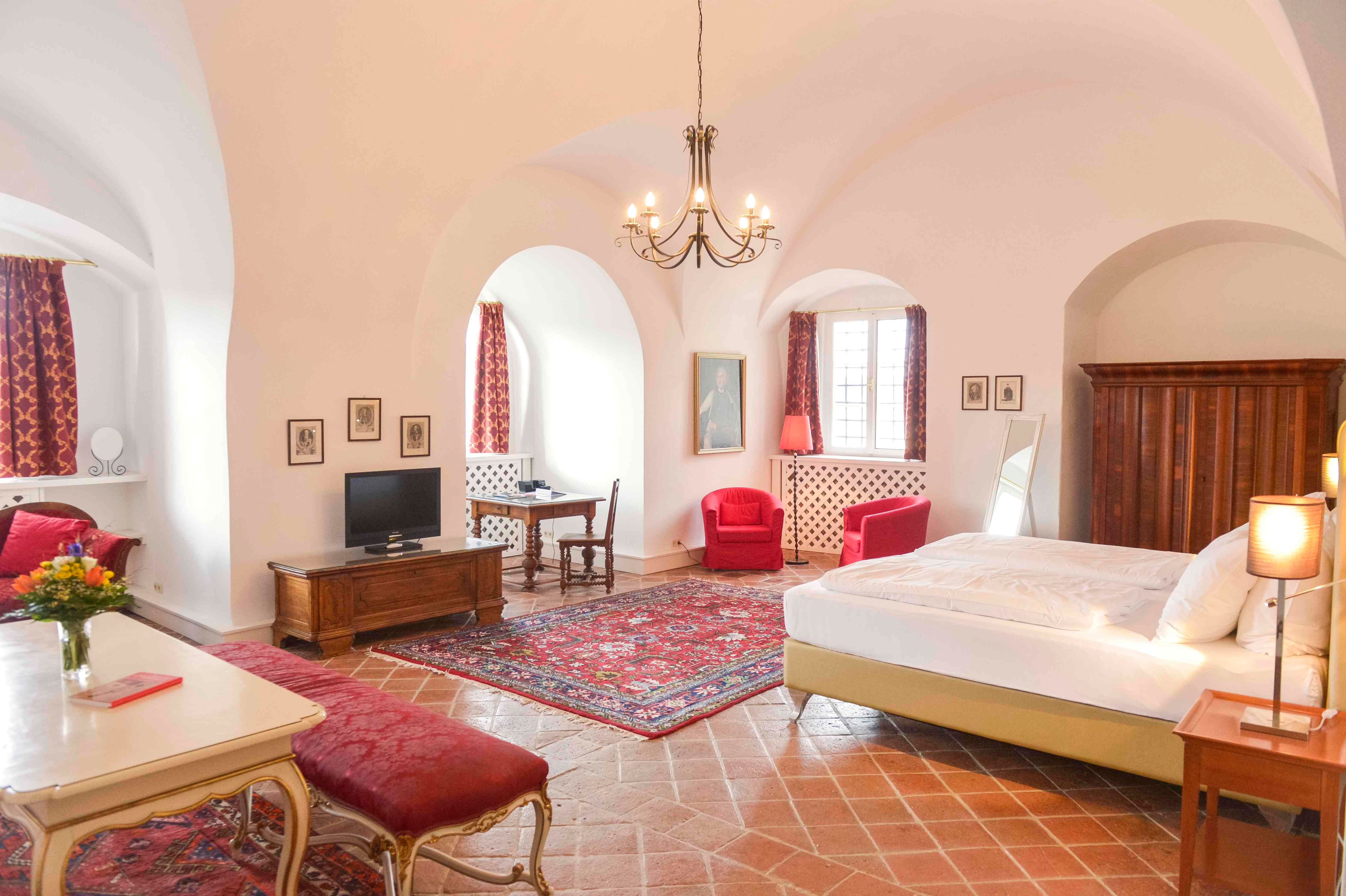 Luxusní apartmá se starožitným nábytkem, červenými koberci a závěsy, velkou postelí a lustrem.