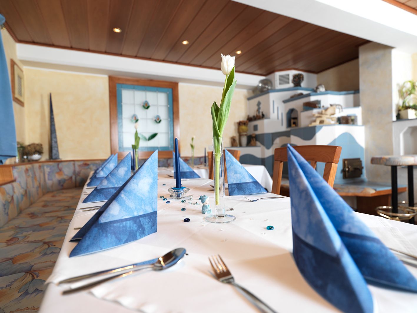 Elegantně prostřený stůl s modrými ubrousky a bílým tulipánem ve sklenici v útulné místnosti.