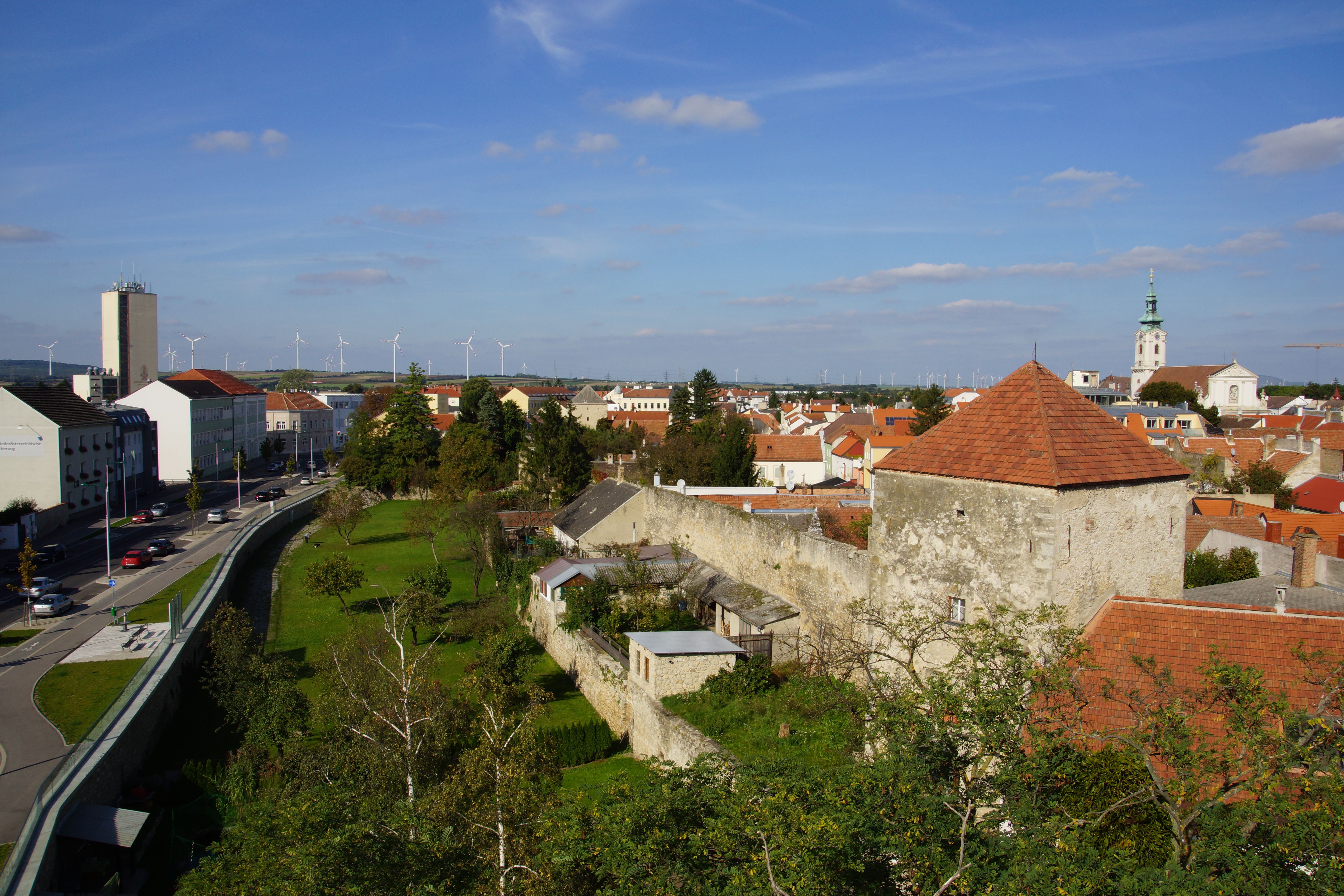 Pohled na město Bruck an der Leitha s městskými hradbami, kostelem a větrnými turbínami v pozadí.