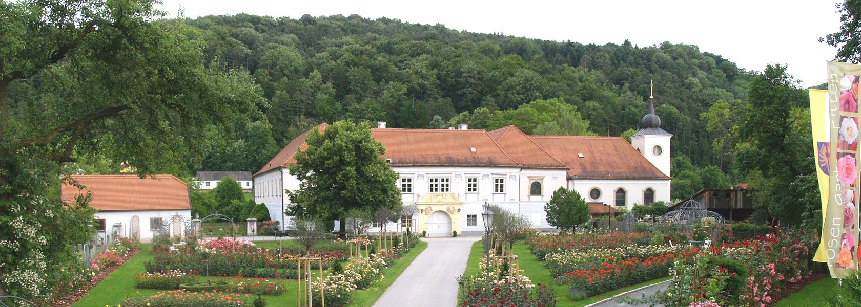 Historická budova s červenou střechou, obklopená růžovou zahradou a stromy, se zalesněným kopcem v pozadí.