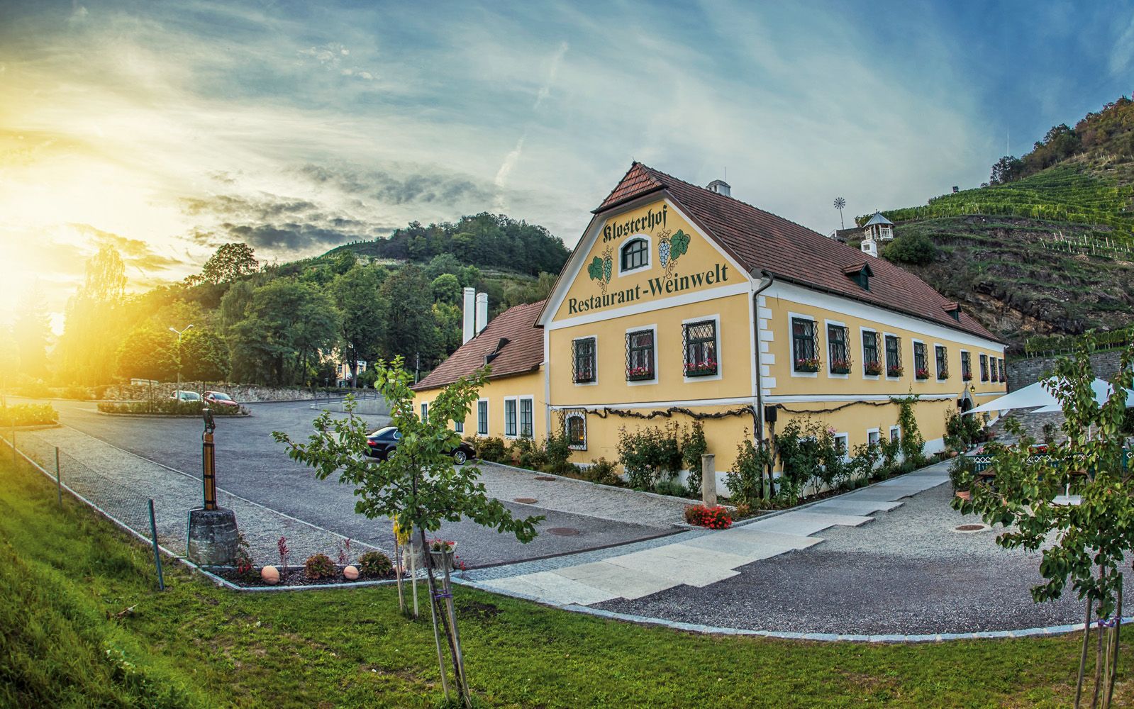 Vnější pohled na žlutou budovu s nápisem "Klosterhof Restaurant-Weinwelt", obklopenou stromy a vinicemi při západu slunce.