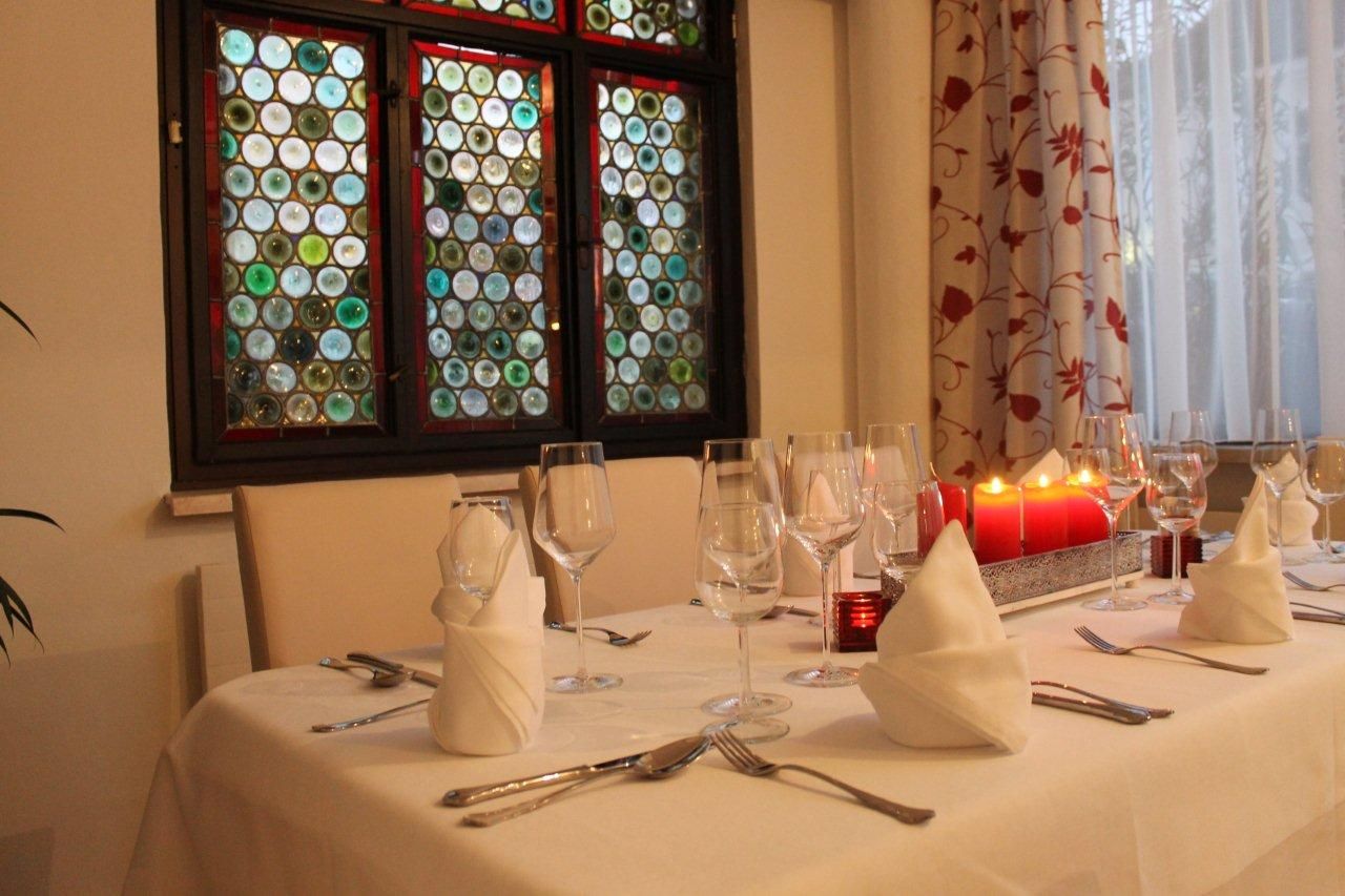 Elegantní stůl s bílými ubrusy, skleničkami na víno a červenými svíčkami před barevným oknem.