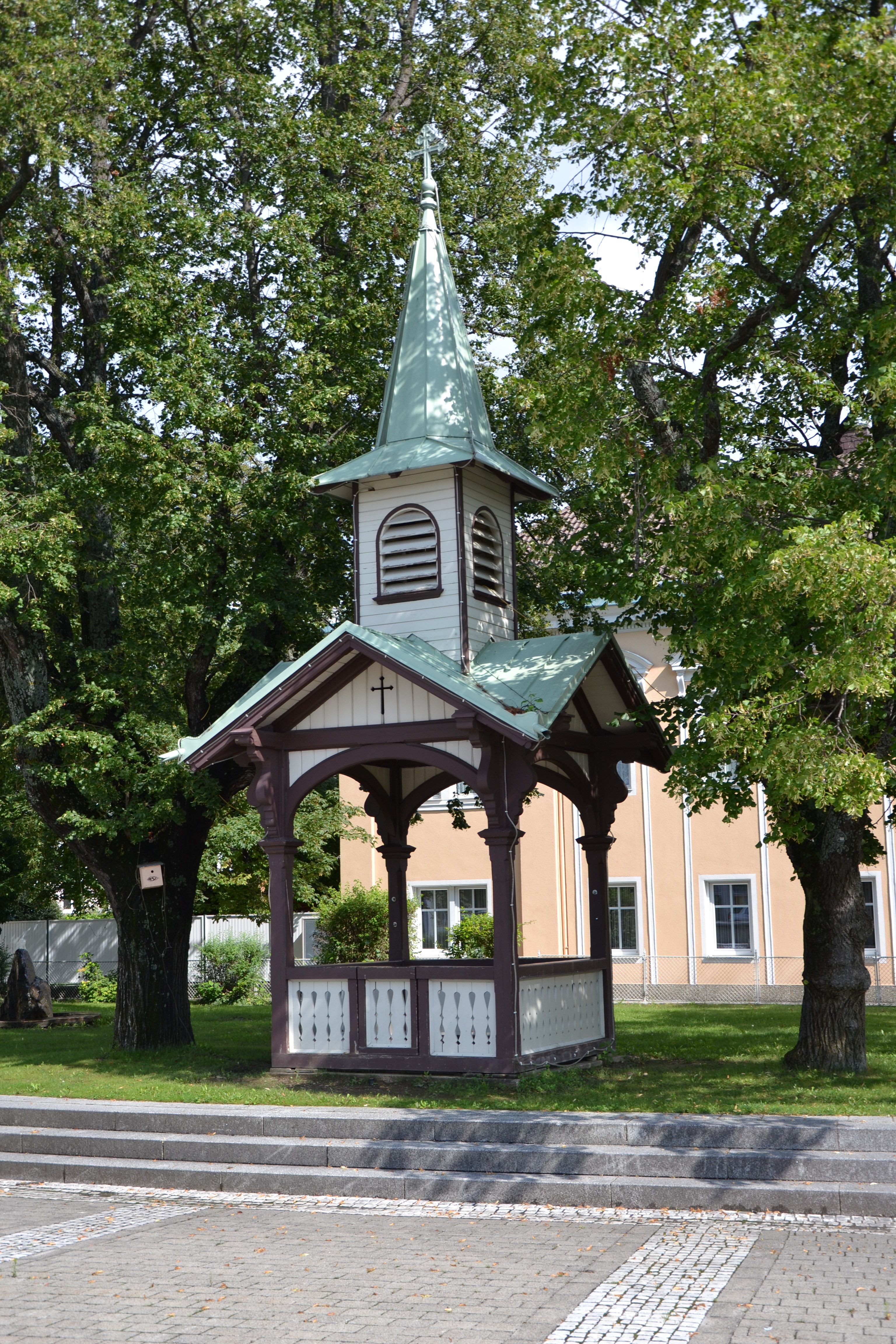 Malý pavilon se špičatou střechou a křížem, obklopený stromy.