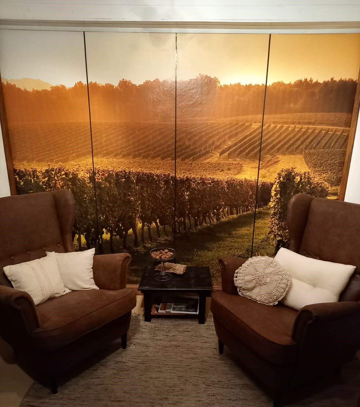 Dvě hnědá křesla s polštáři před stěnou s krajinnou fototapetou vinice.
