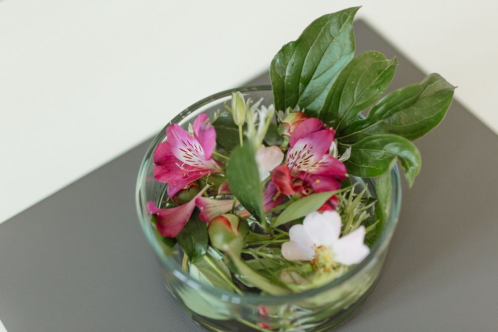 Skleněná nádoba s barevnými květy a zelenými listy na stole.