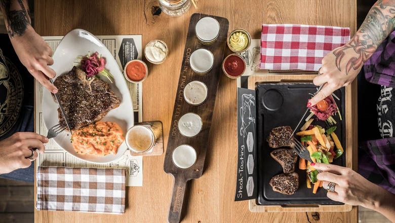 Dvě osoby jedí steaky s přílohami u dřevěného stolu.