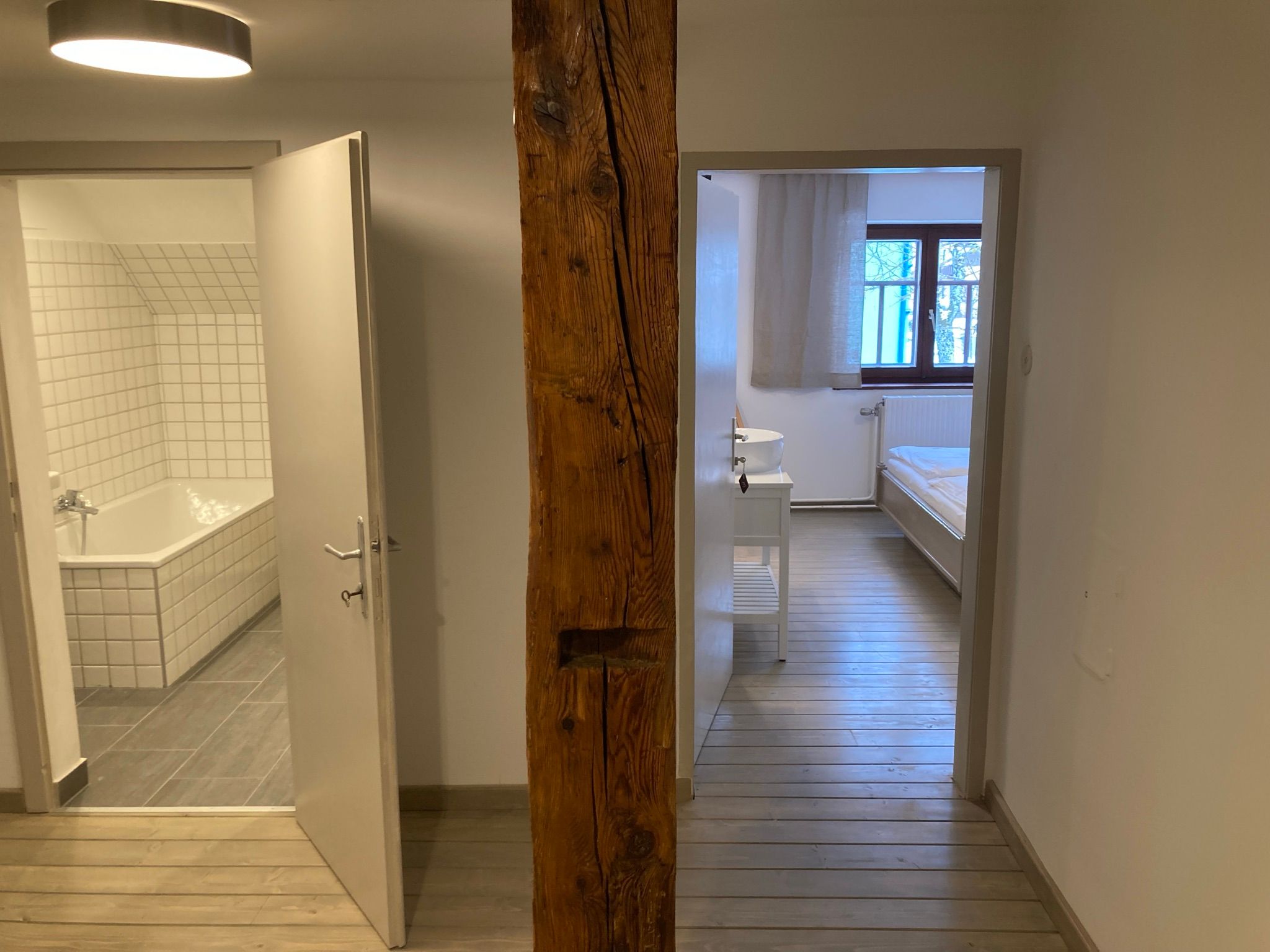 Jedna koupelna a jedna ložnice s dřevěnou podlahou, oddělená dřevěnou trámovou stěnou.