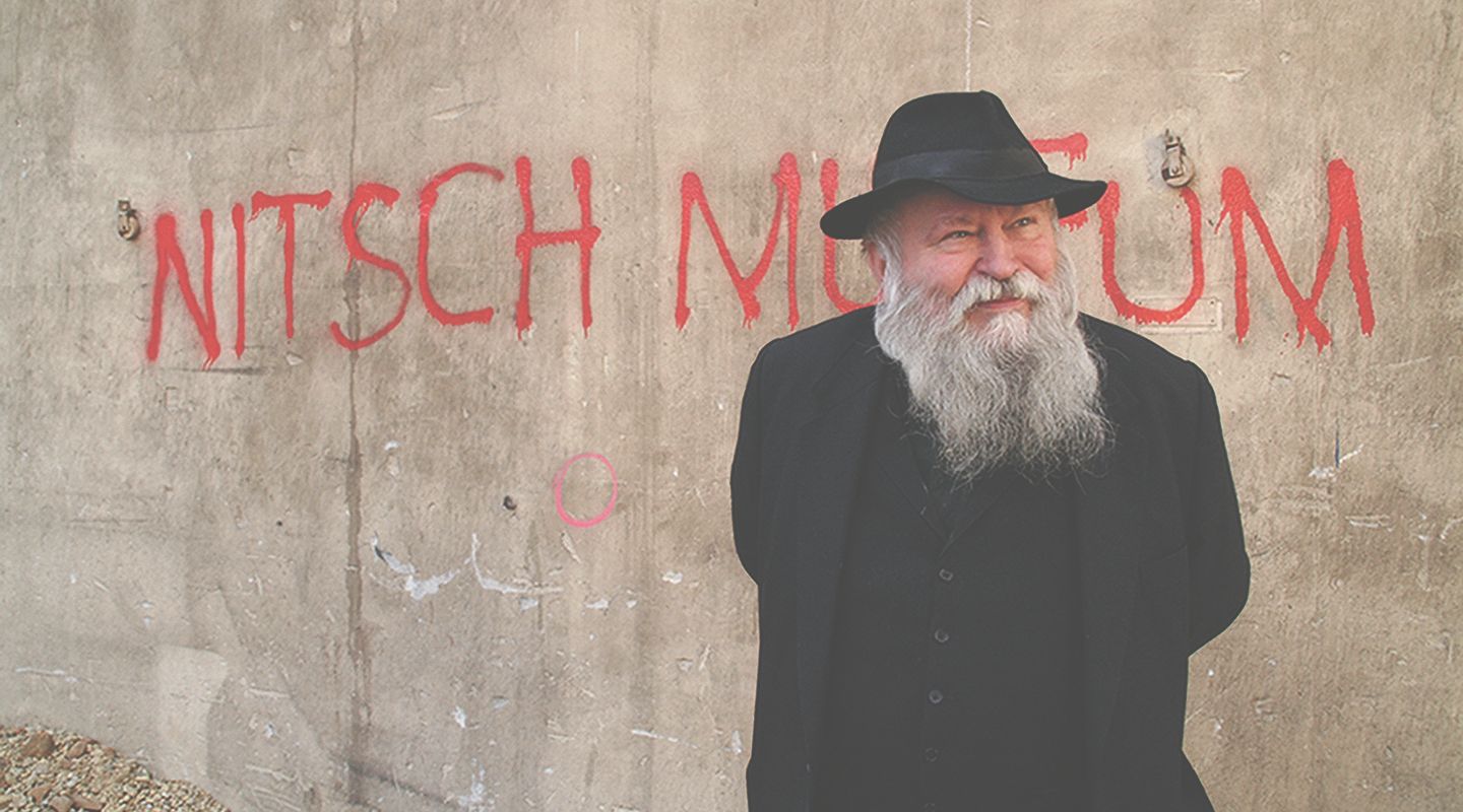 Muž s plnovousem a kloboukem stojí před stěnou s nápisem "Nitschovo muzeum".