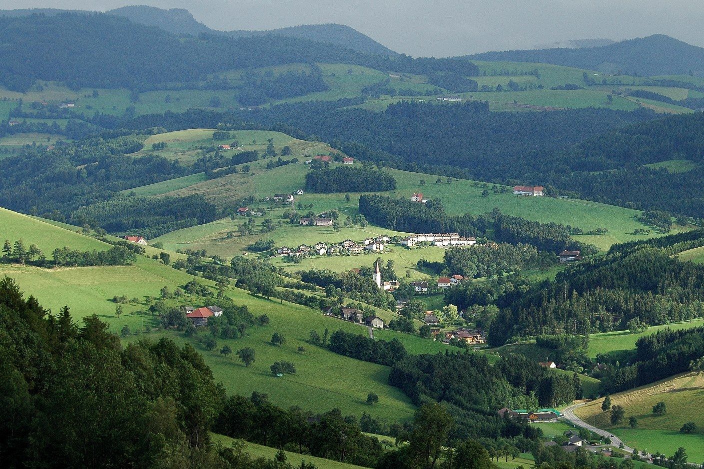 Panoramatický pohled na zelenou kopcovitou krajinu s roztroušenými domy a lesy.