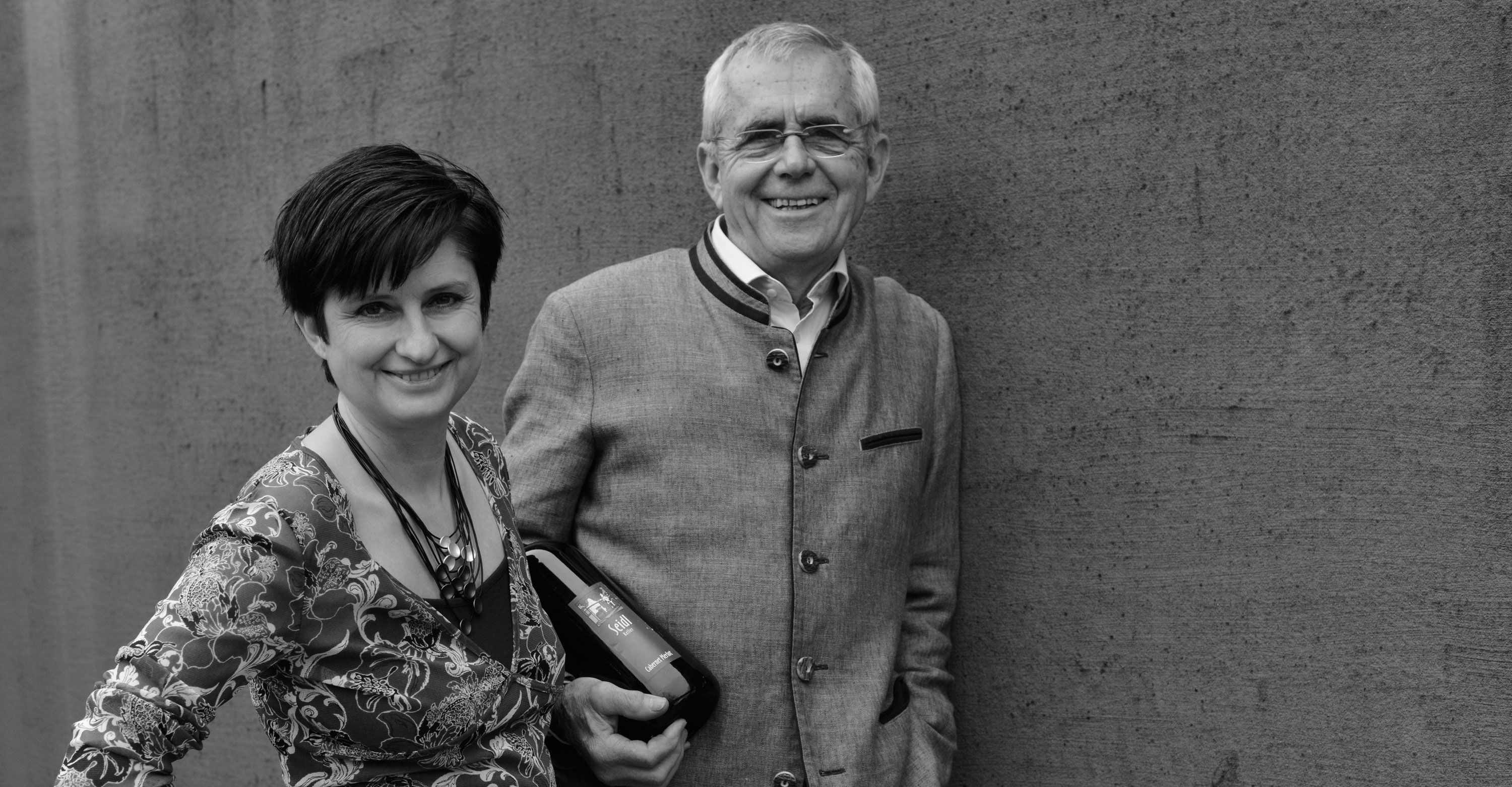 Muž a žena stojí s úsměvem před zdí. Muž drží láhev vína.