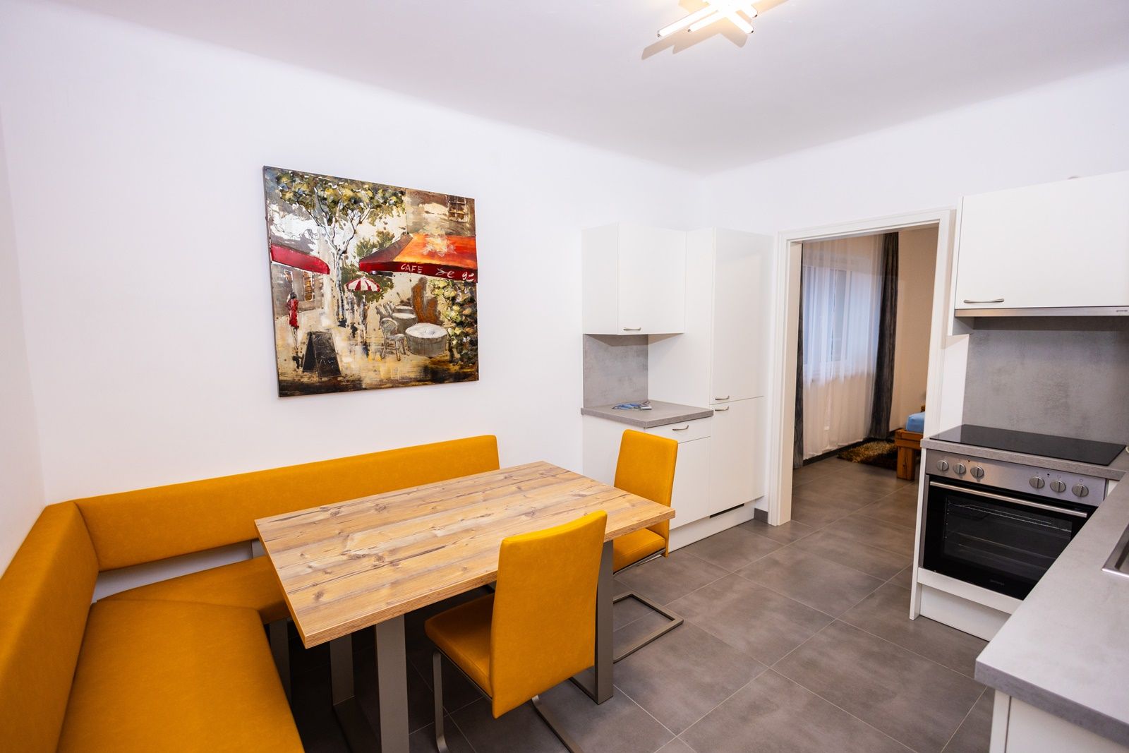 Moderní kuchyně se žlutou rohovou lavicí, dřevěným stolem a uměleckými díly na stěně.