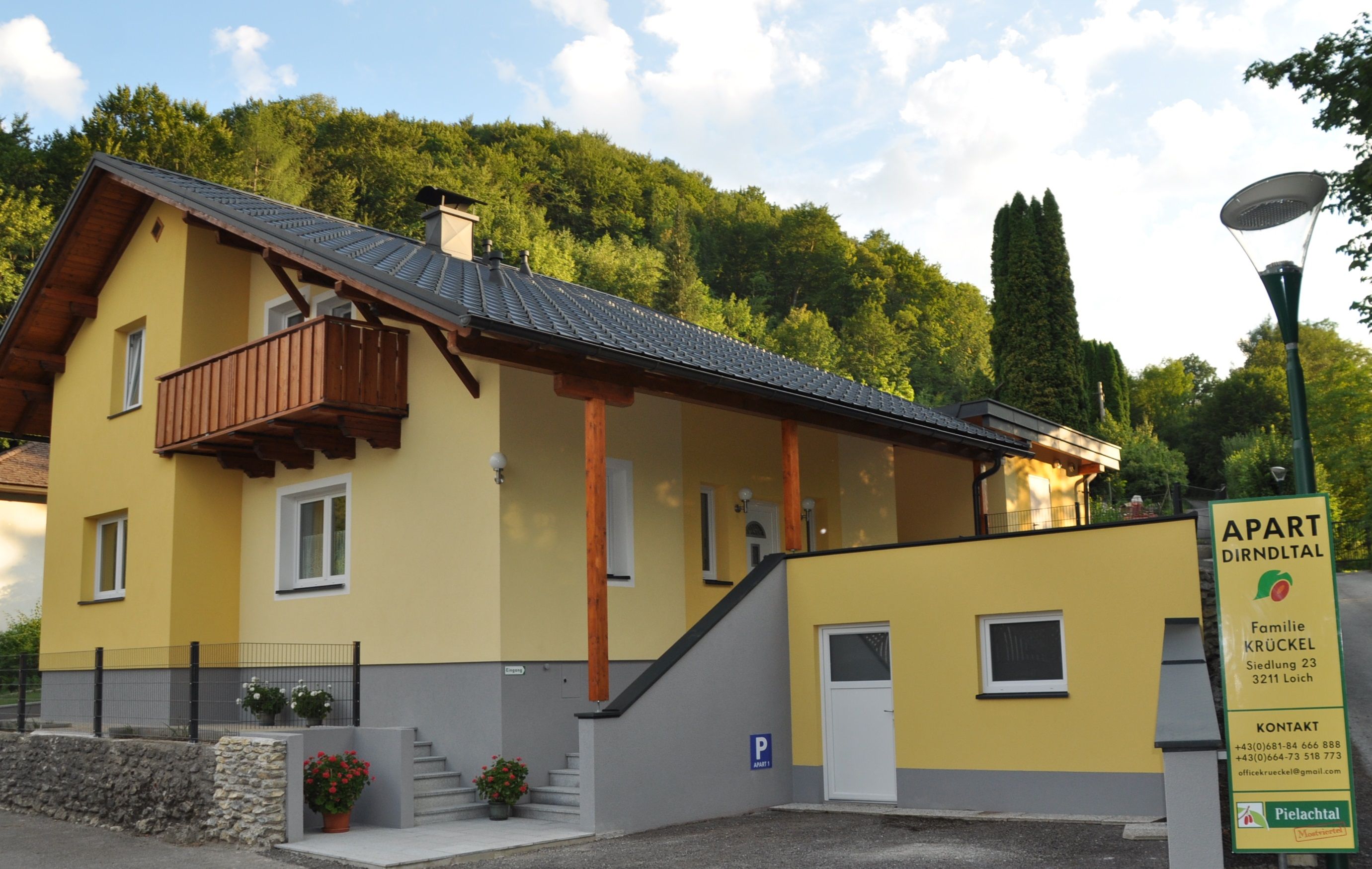 Žlutý dům s balkonem a nápisem "Apart Dirndltal" před zalesněným kopcem.