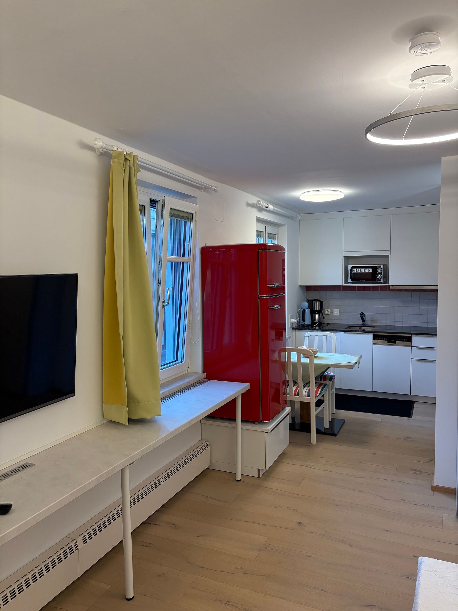 Moderní obývací pokoj s otevřenou kuchyní, červenou lednicí, žlutými závěsy a dřevěnou podlahou.