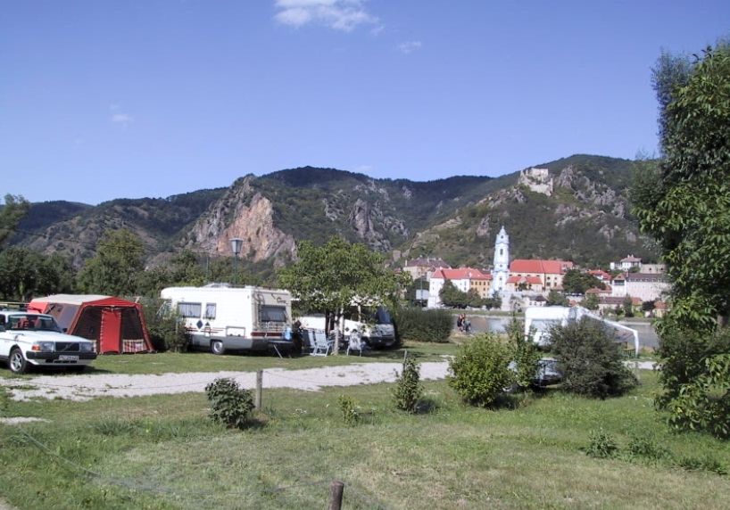 Kemp s karavany a stany před horskou krajinou a kostelem v pozadí.