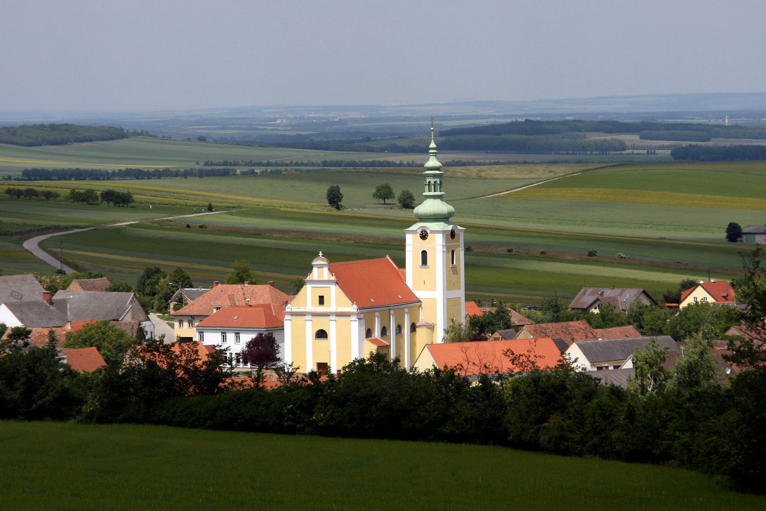 Pohled na kostel v Ottenthalu s okolními domy a rozlehlými poli v pozadí.