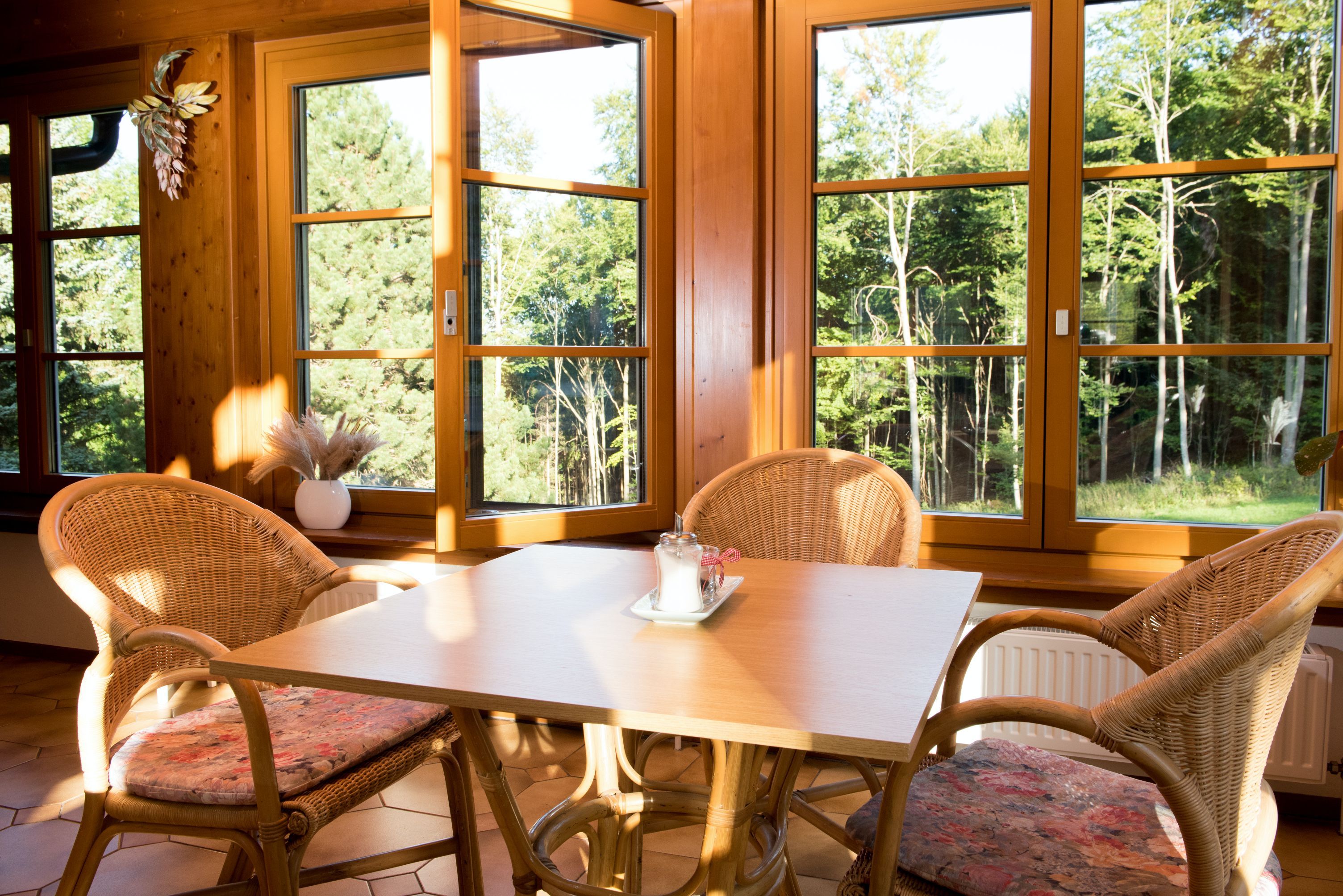 Stůl se čtyřmi ratanovými židlemi ve slunné místnosti s velkými okny a výhledem na les.