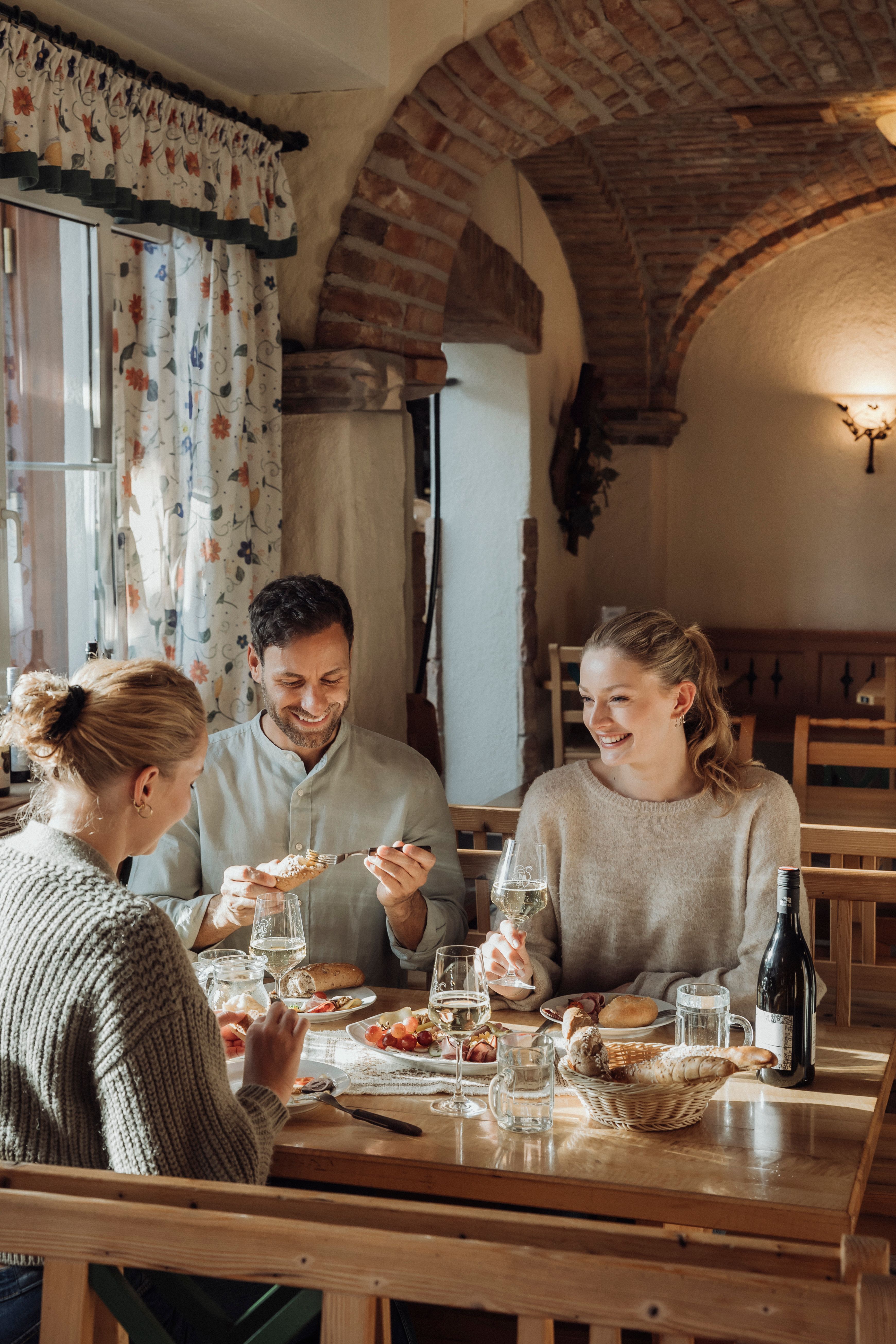 Tři lidé sedí u stolu v útulné restauraci a vychutnávají si víno a jídlo.