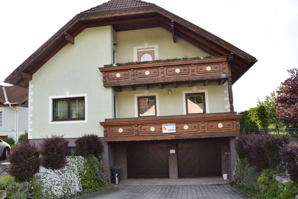 Dvoupodlažní dům se zelenou fasádou a dřevěnými balkony, obklopený upravenými zahradami.