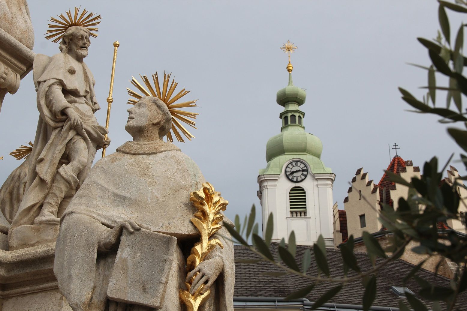 Socha se svatozáří před kostelní věží s hodinami a zelenou střechou.
