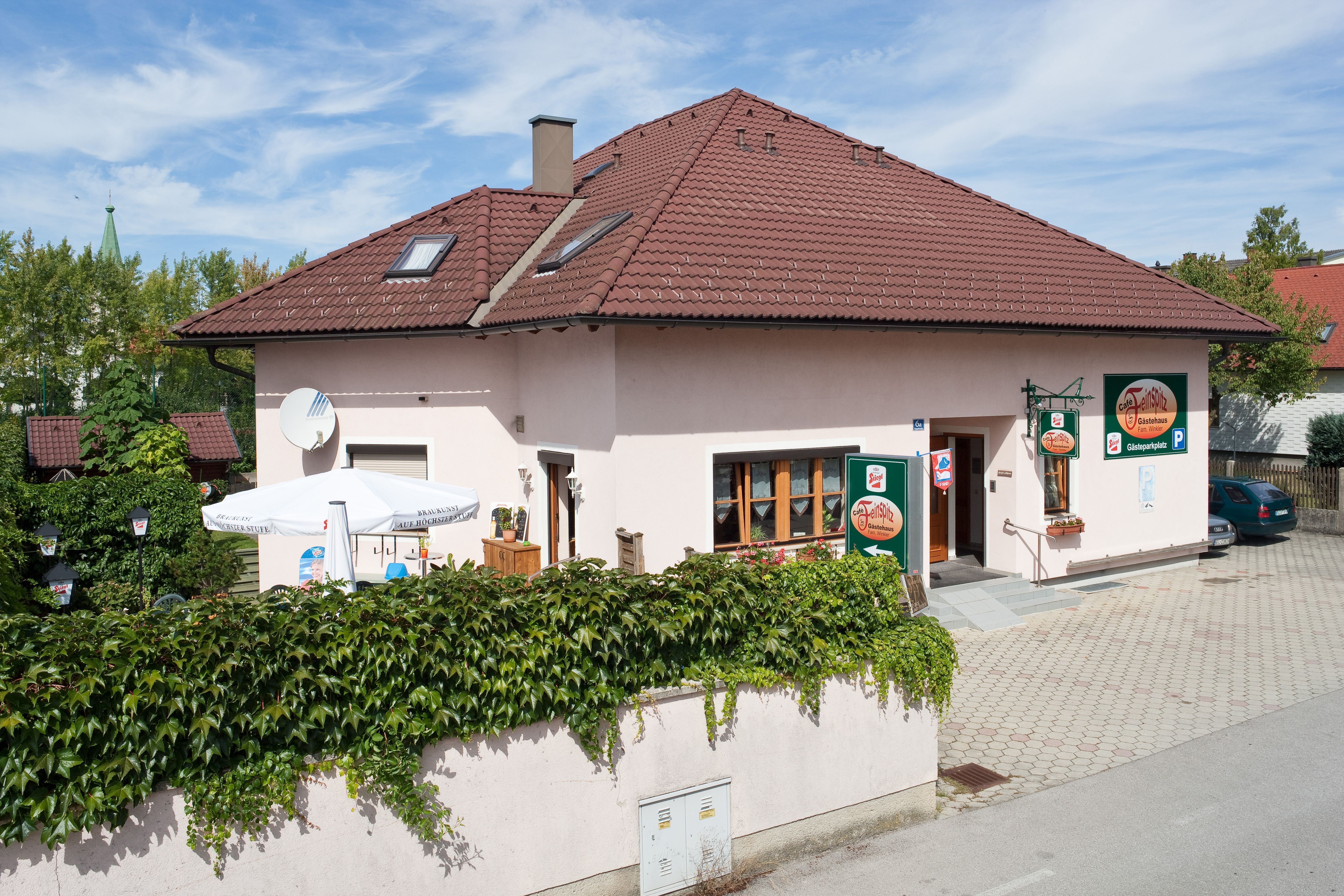 Penzion s červenou střechou a terasou, obklopený zelení.