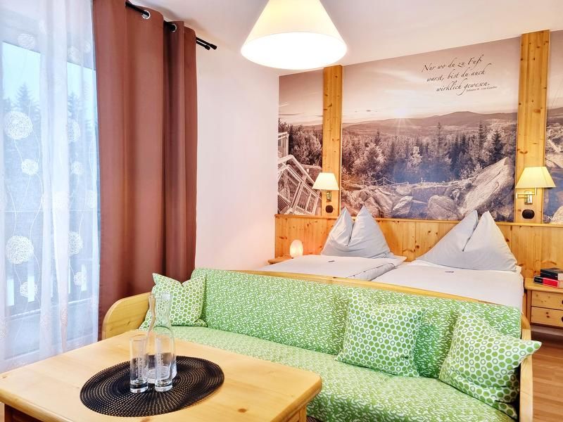 Útulný dvoulůžkový pokoj se dvěma lůžky, zelenou pohovkou a velkou nástěnnou malbou krajiny.