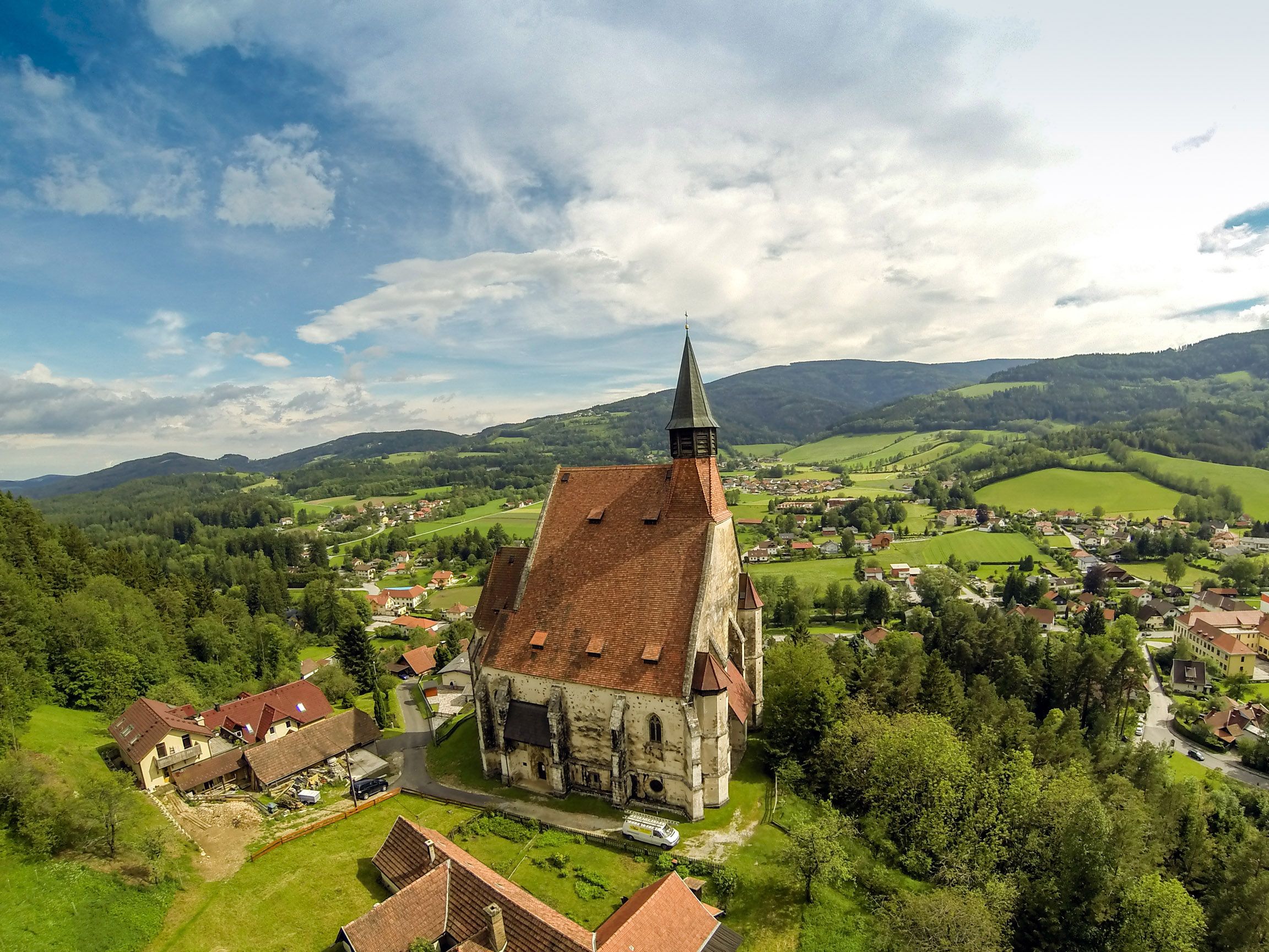 Letecký pohled na kostel ve venkovském prostředí s kopci a lesy v pozadí.
