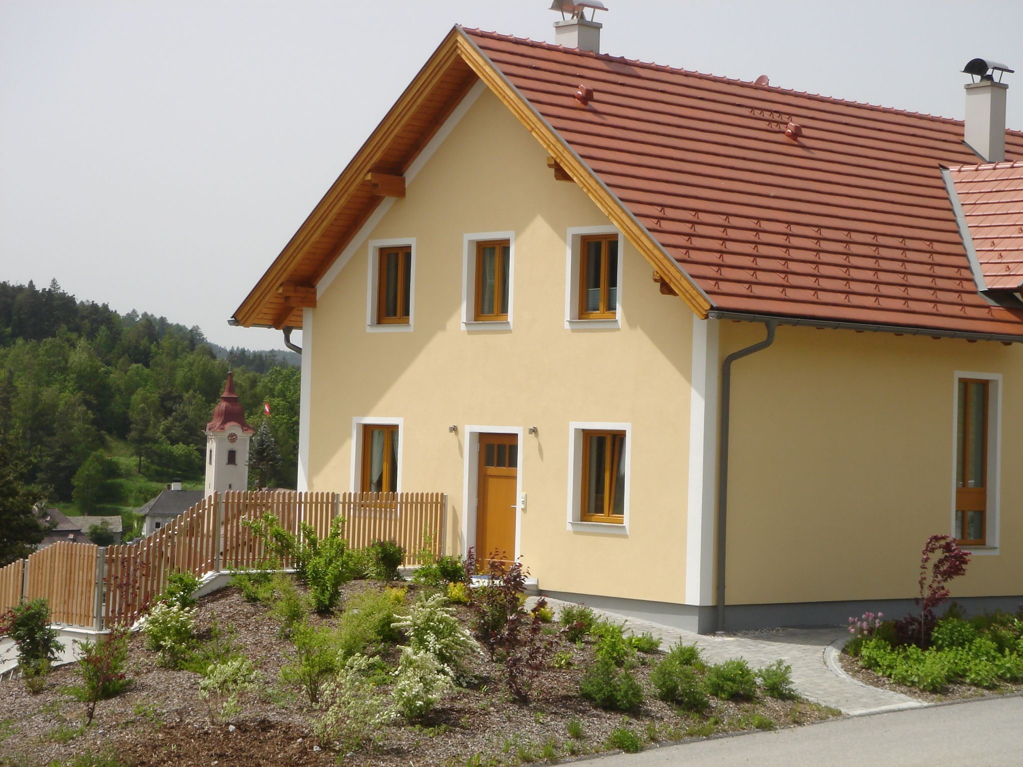 Moderní žlutý dům s červenou střechou a zahradou, v pozadí kostel a zelená krajina.