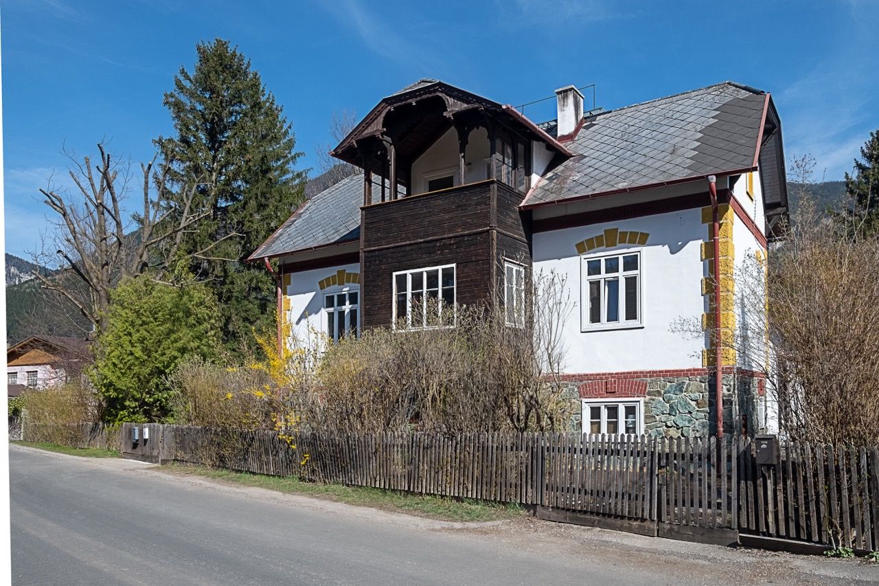 Tradiční venkovský dům s dřevěným balkonem a bílou fasádou, obklopený stromy a keři, pod jasně modrou oblohou.