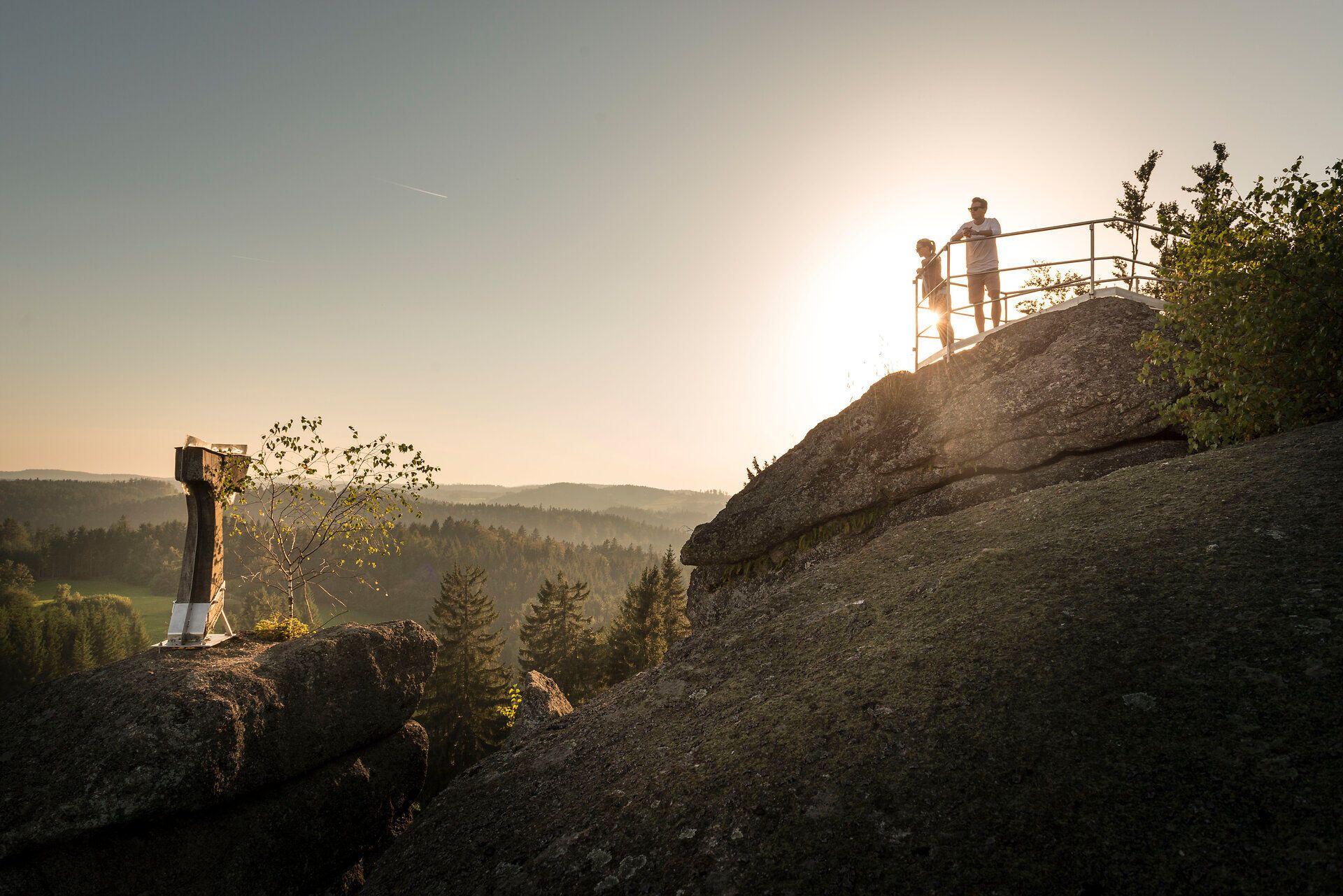 Zlaté večerní slunce zalévá majestátní skály hřejivým světlem a dva turisté si užívají úchvatný výhled. Toto místo obklopené panenskou přírodou a jemným šuměním stromů vybízí k odpočinku a poznávání krásy hor.