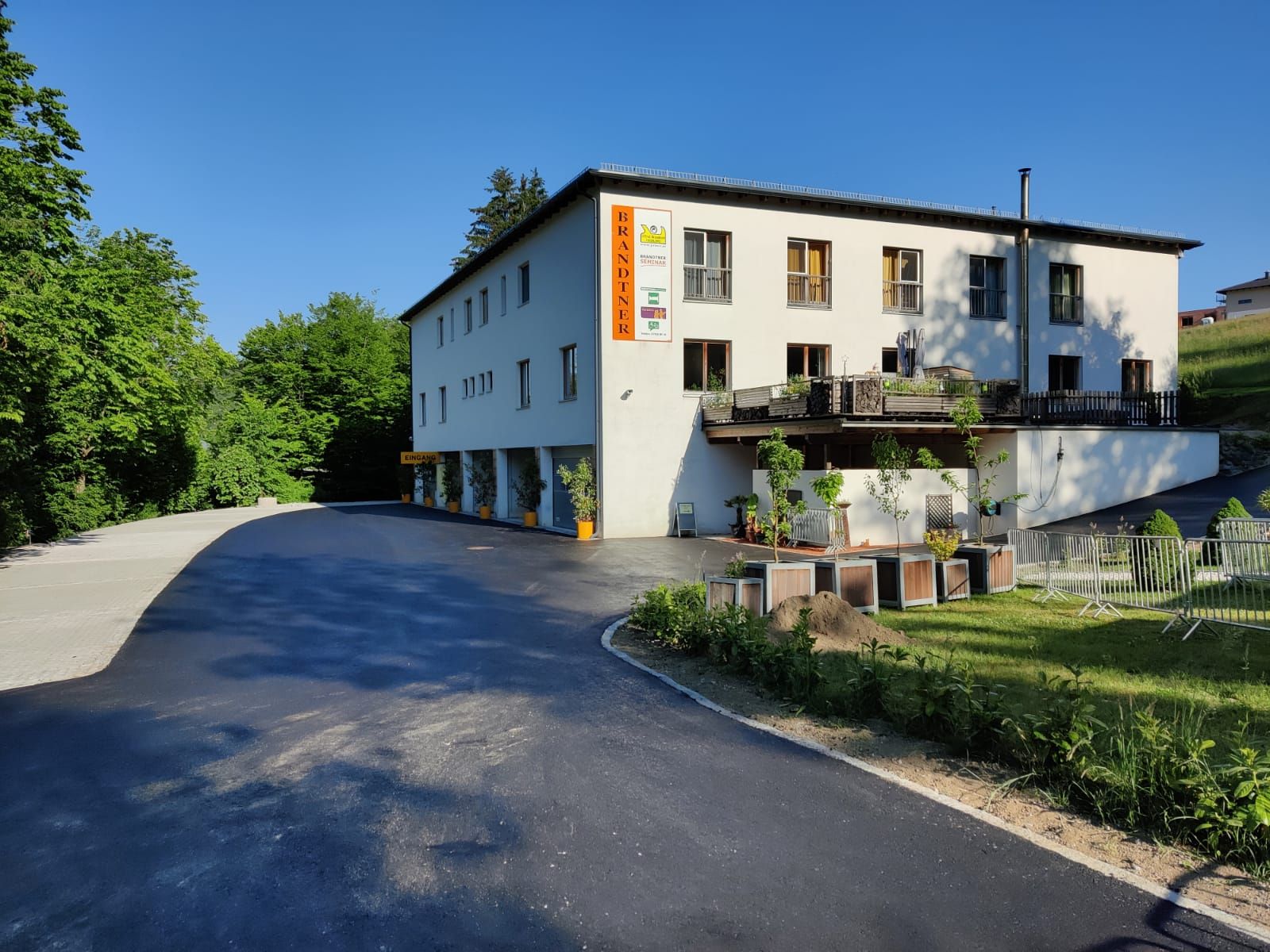 Budova s nápisem "Brandtner", obklopená stromy a parkovištěm.