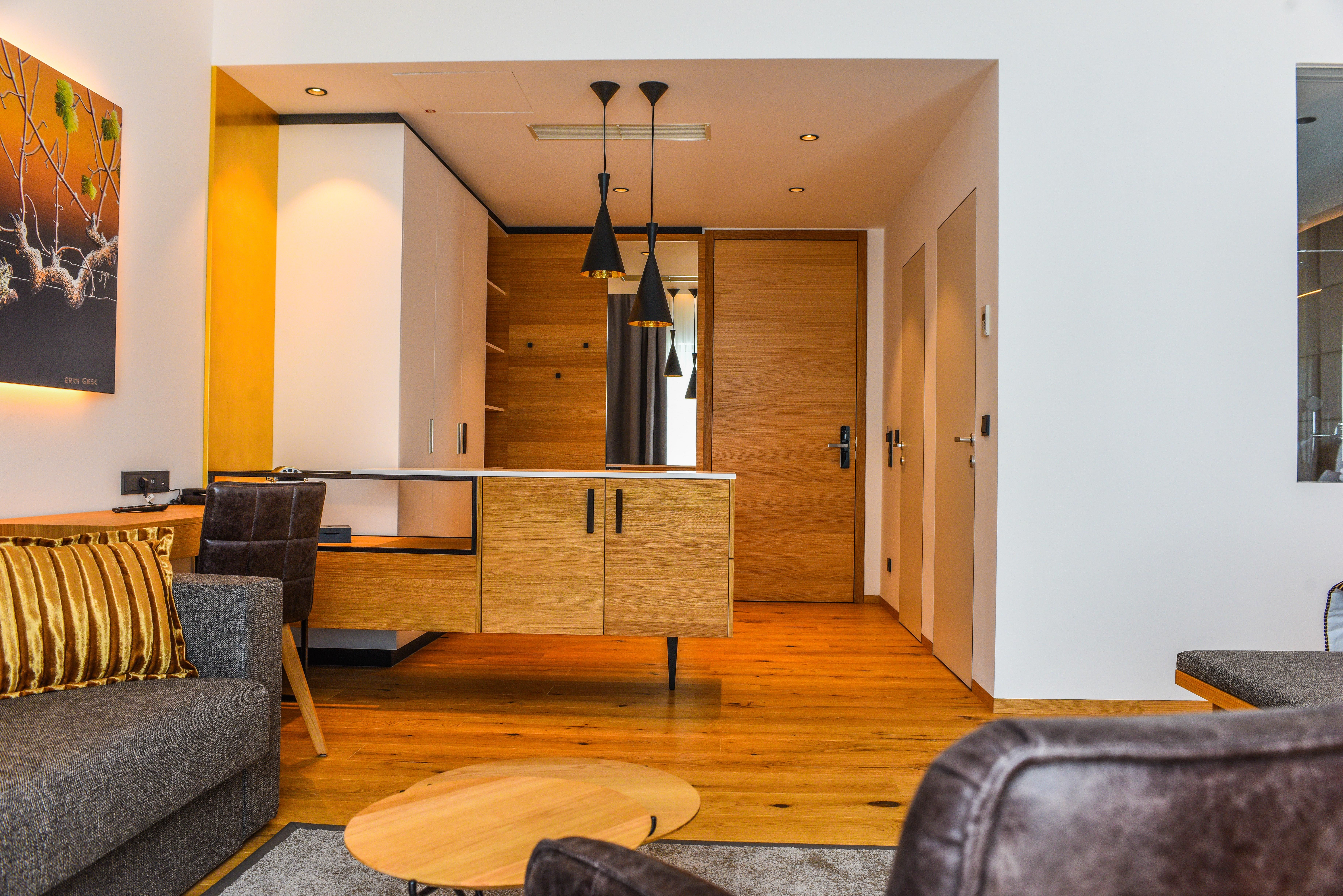 Pohled do interiéru moderního hotelového apartmá s dřevěnou podlahou, šedou pohovkou a dřevěným psacím stolem.