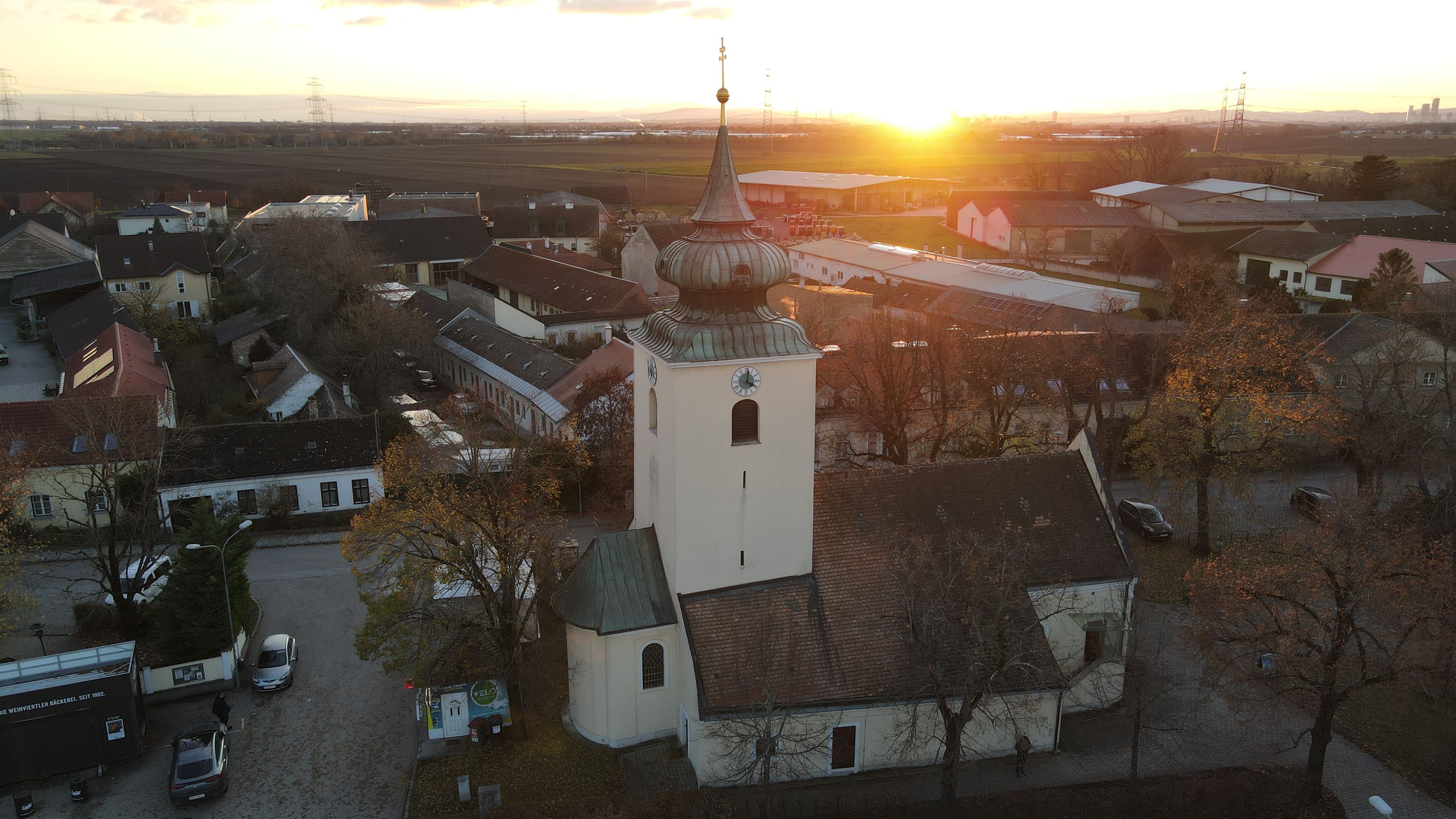 Kostel v Raasdorfu s věží a cibulovitou střechou při západu slunce, obklopený domy a poli.