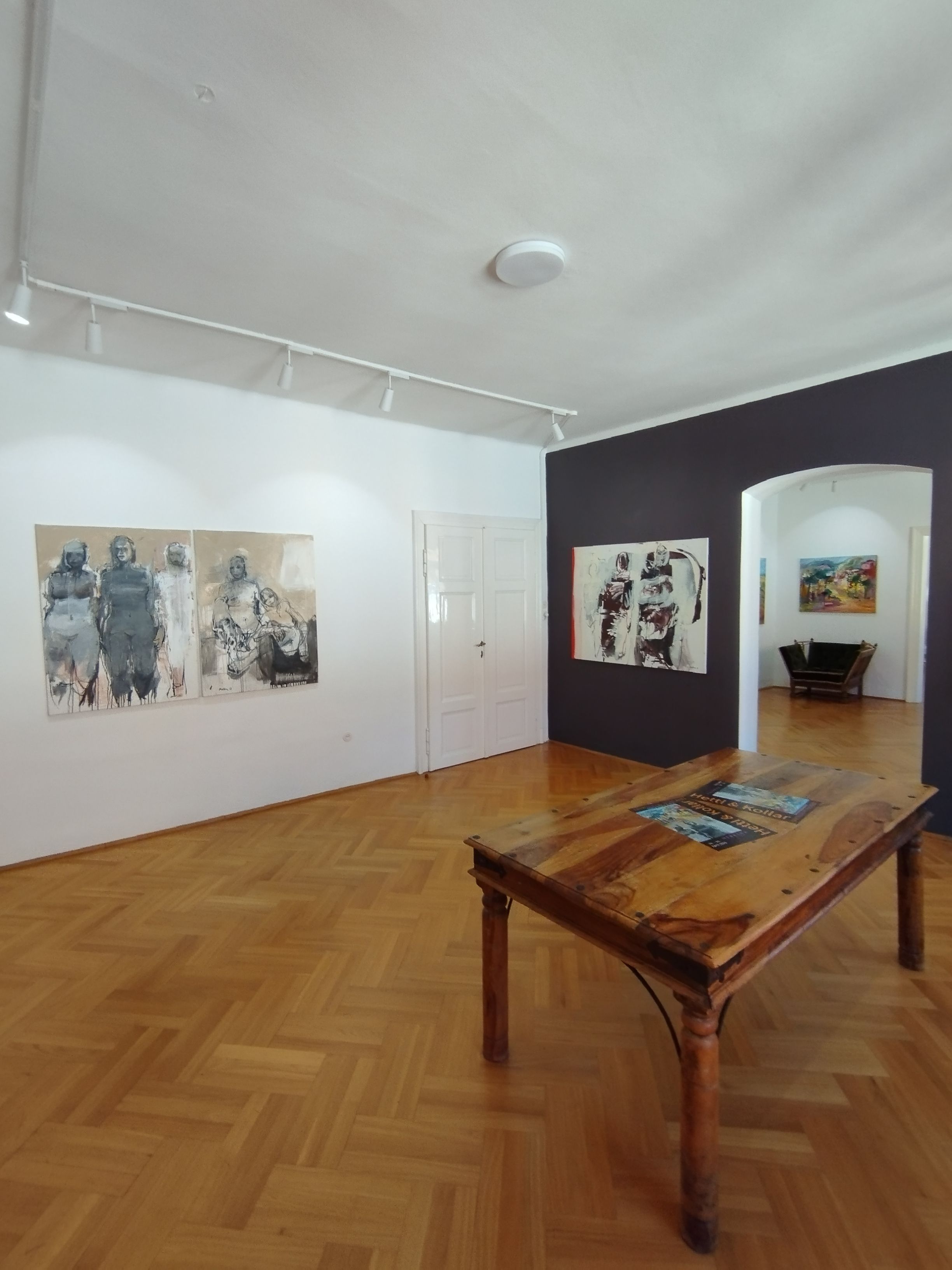 Pohled do interiéru umělecké galerie s dřevěnou podlahou, obrazy na stěnách a dřevěným stolem uprostřed.