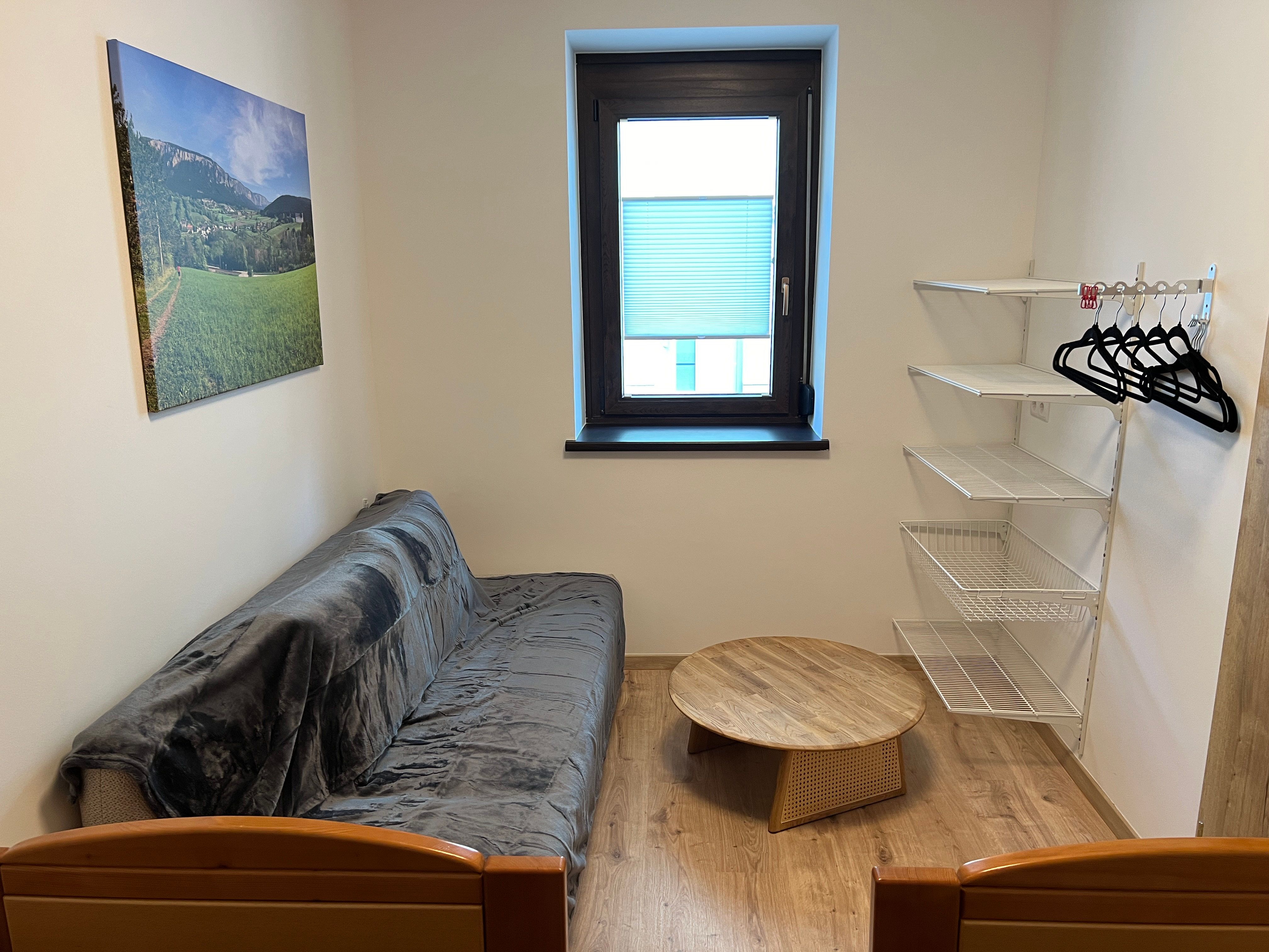 Malý pokoj s pohovkou, kulatým dřevěným stolem, nástěnnou policí a oknem.