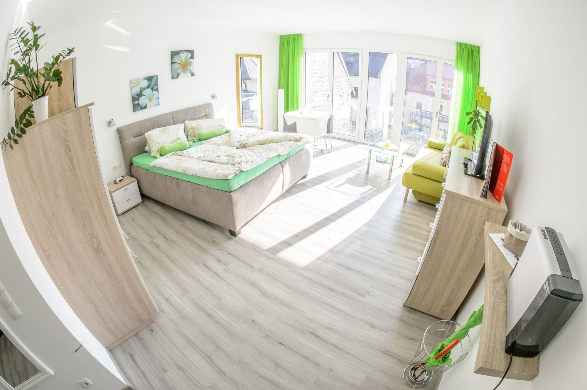 Světlý pokoj pro hosty s manželskou postelí, pohovkou, TV a velkými okny.