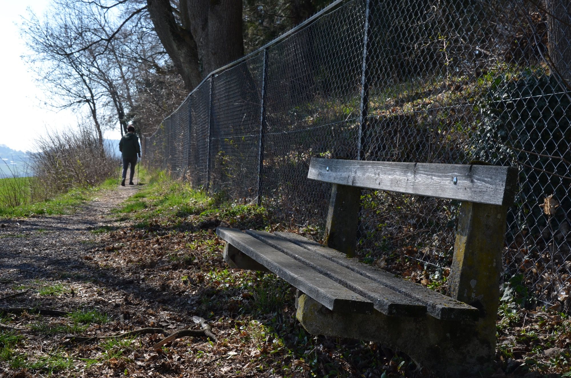 Chodník s dřevěnou lavičkou a plotem z drátěného pletiva. V dálce kráčí člověk.