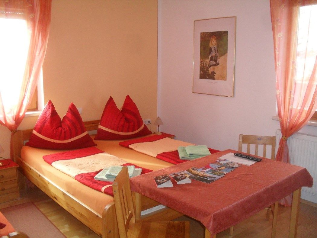 Útulná ložnice s manželskou postelí, červenými polštáři a stolkem s brožurami.