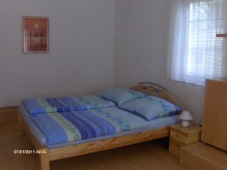 Jednoduše zařízená ložnice s manželskou postelí, nočním stolkem a oknem se závěsy.