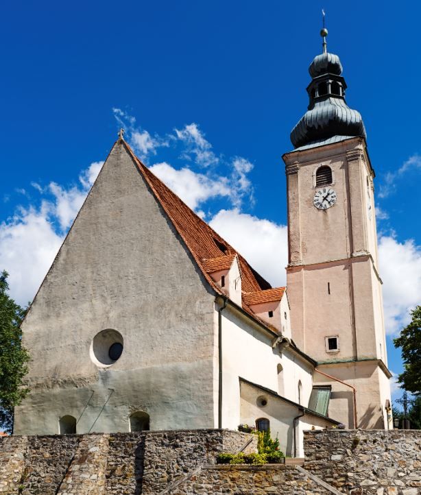 Farní kostel Wiesmath s věží, kamennou hřbitovní zdí a hodinami na pozadí modré oblohy.
