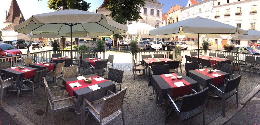 Venkovní terasa kavárny se stoly a slunečníky na dlážděném náměstí.