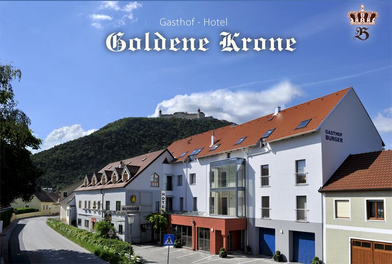 Gasthof-Hotel Goldene Krone před zalesněným kopcem s hradem v pozadí.