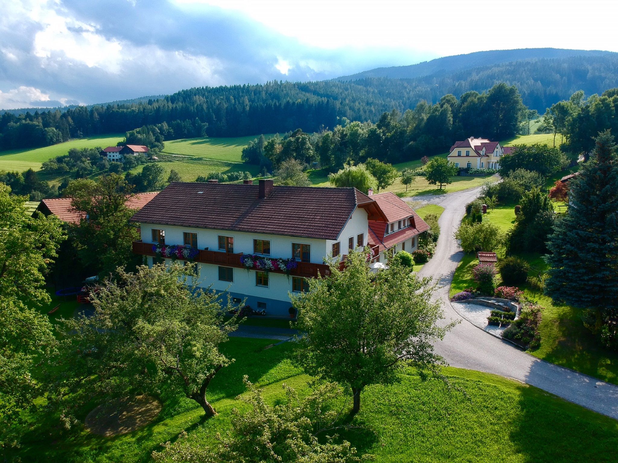 Venkovská krajina s velkým domem, obklopená stromy a loukami, pod zataženou oblohou.