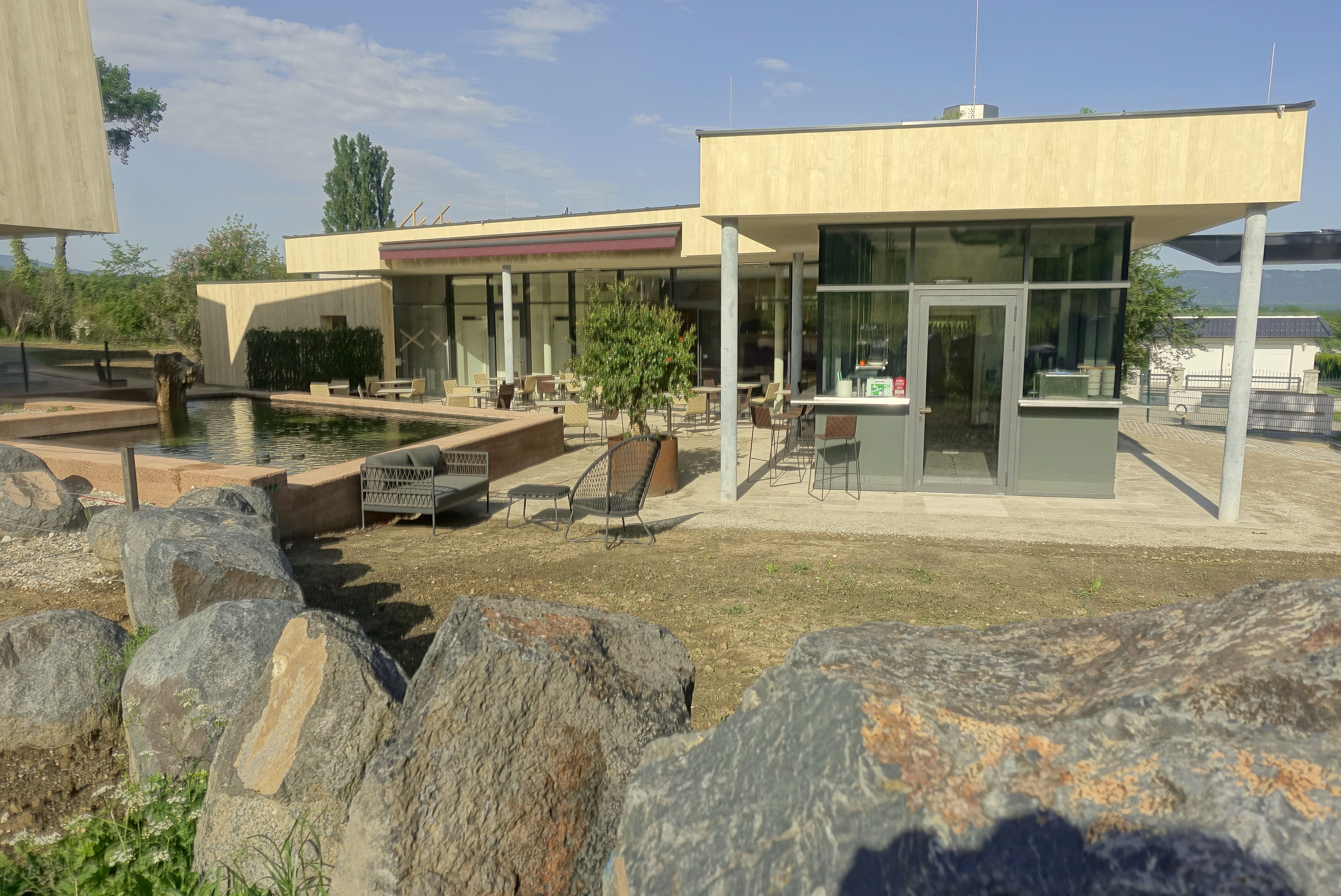 Moderní veranda s proskleným průčelím a posezením, obklopená skalami a jezírkem.