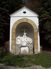 Malá kaple s křížem a náboženskými postavami ve výklenku.
