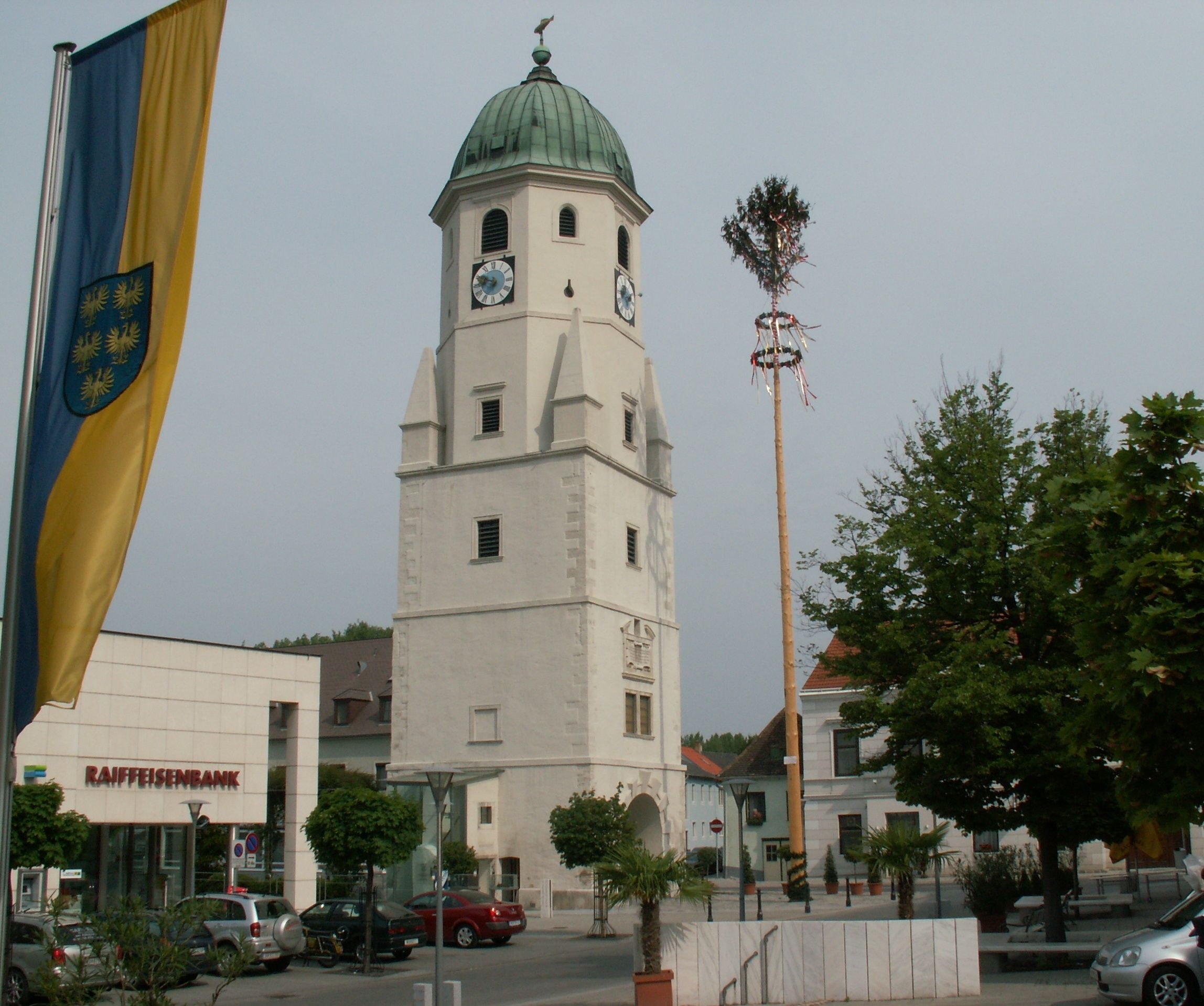 Kostelní věž s hodinami a zelenou střechou ve Fischamendu, vedle májky a pobočky Raiffeisenbank.