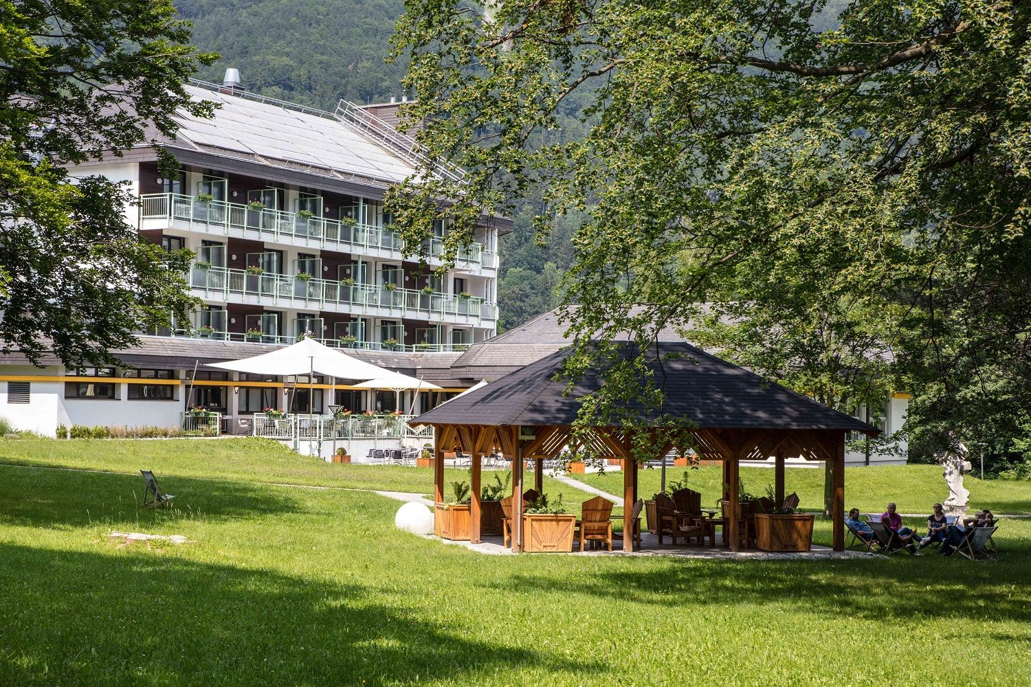 Hotelová budova s balkony v pozadí, pavilon a lidé na židlích v popředí, obklopené stromy a trávníky.