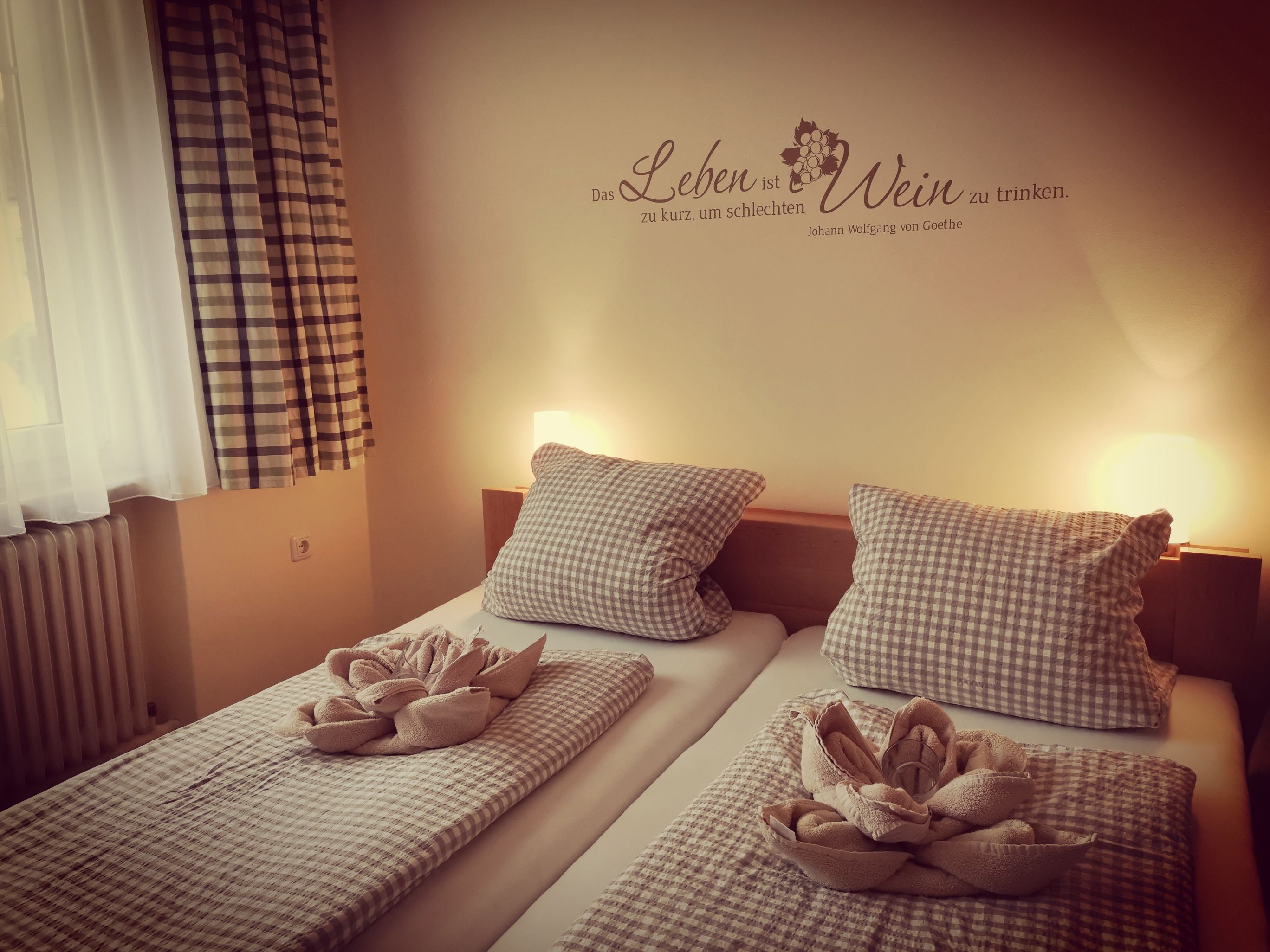 Útulný pokoj pro hosty se dvěma postelemi, kostkovanými přikrývkami a polštáři, ozdobnými ručníky a Goethovým citátem na zdi.