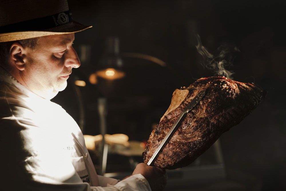 Muž v bílé košili drží nožem velký grilovaný kus masa.